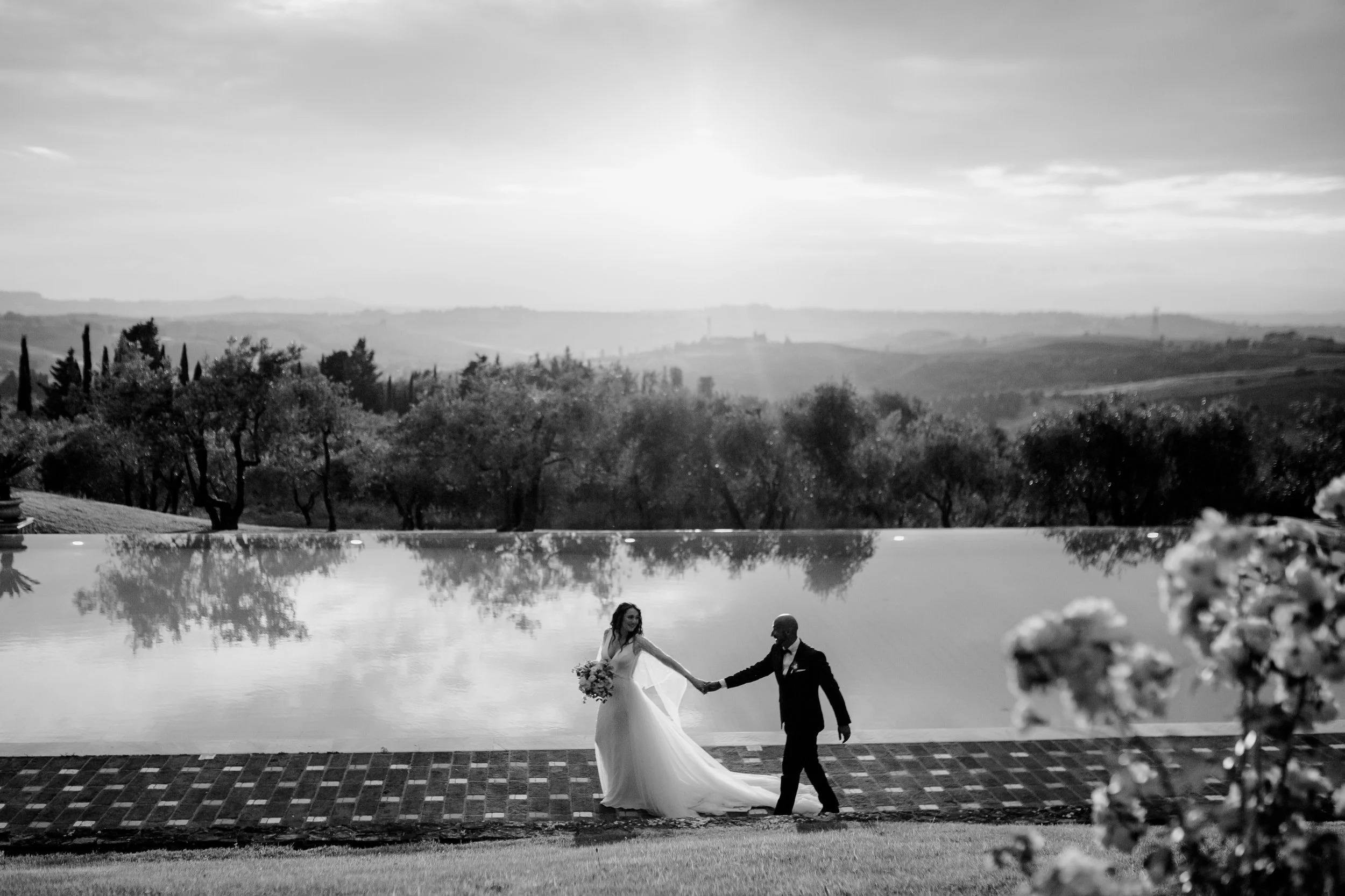 Una coppia di sposi cammina vicino a una piscina in un paesaggio collinare al tramonto, il tutto in bianco e nero.