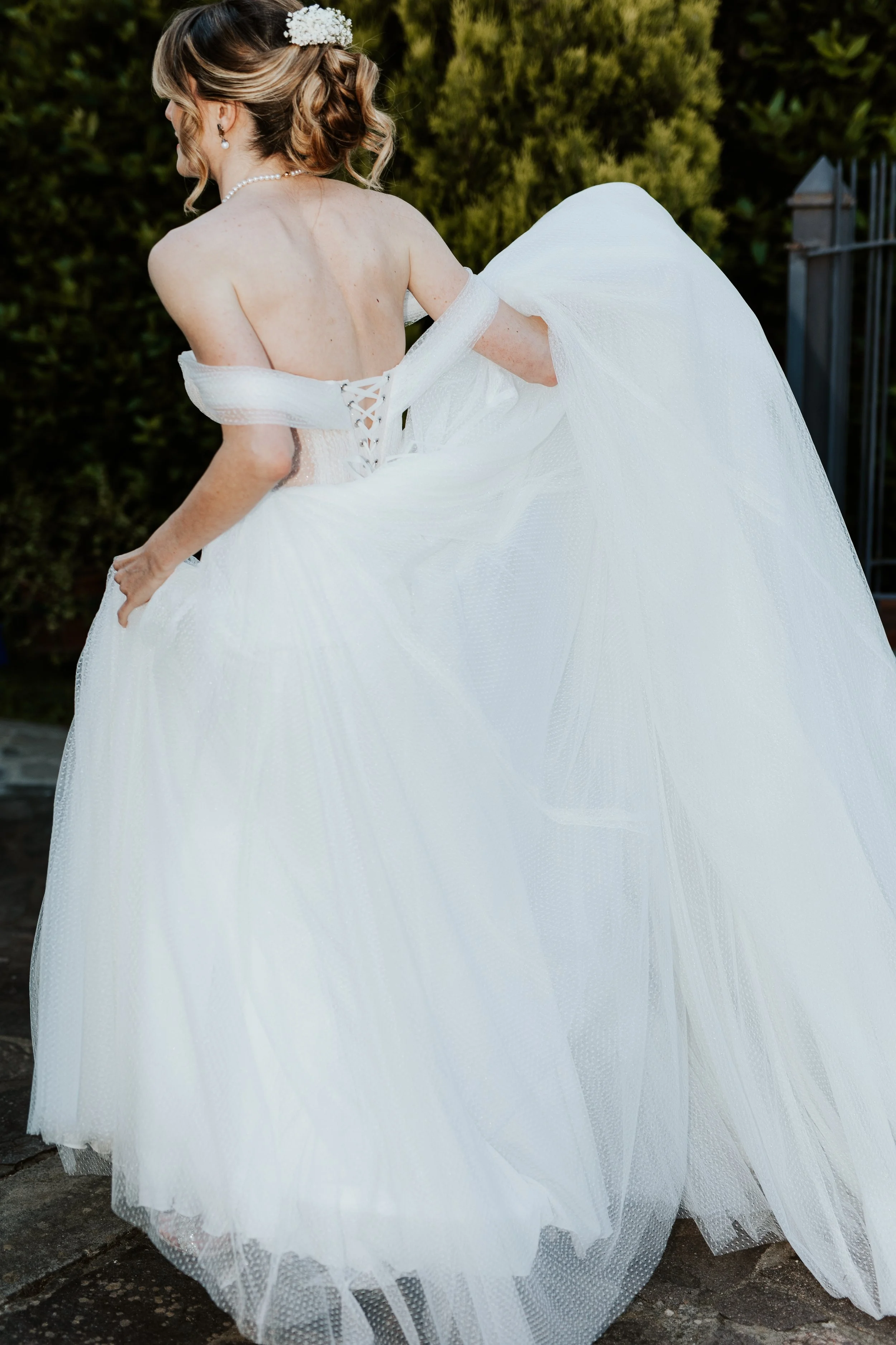 Una donna indossa un abito da sposa bianco con un velo e un'acconciatura con fiori, ritratta di spalle in un giardino.