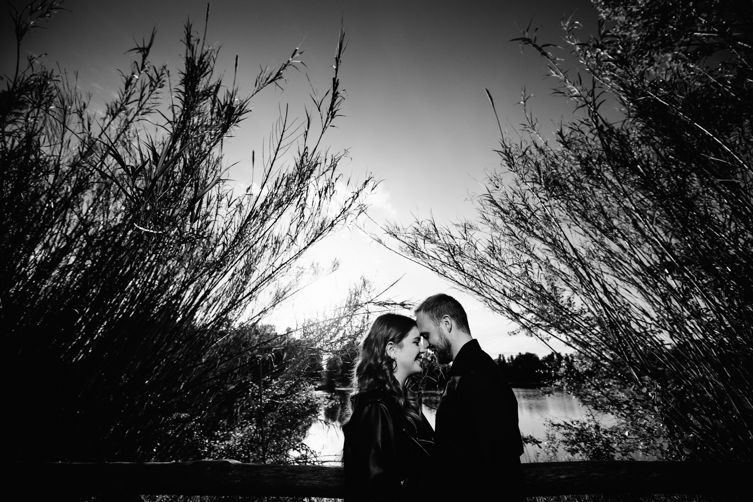 Una coppia romantica si abbraccia e si guarda negli occhi in un paesaggio naturale con cespugli e un fiume, in bianco e nero.
