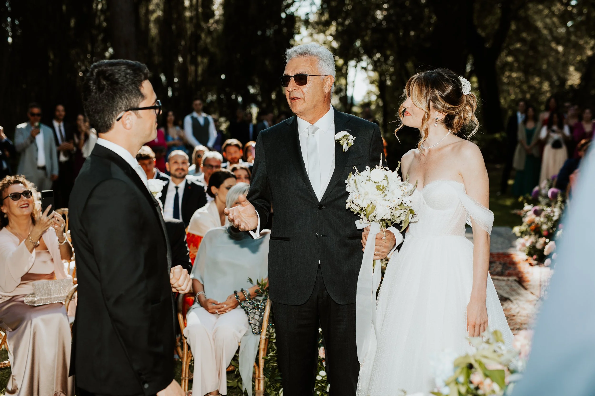 Coppia di sposi al loro matrimonio, con il padre della sposa che parla con lo sposo, circondati da amici e parenti in un ambiente all'aperto.
