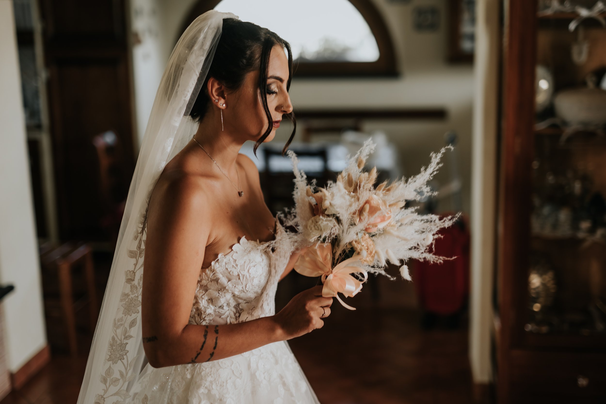 Sposa con abito da matrimonio e velo, che tiene un bouquet di fiori secchi di colore neutro, in una stanza elegante.