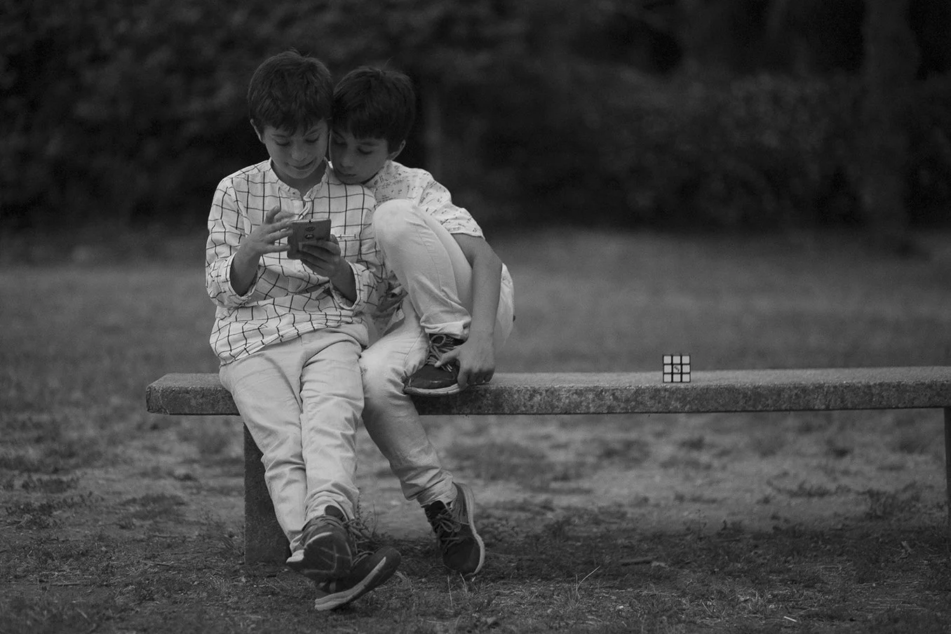 Due bambini seduti su una panchina in un parco, guarda uno di loro sta usando uno smartphone, mentre di fronte a loro c'è un cubo di Rubik posizionato sulla panchina.