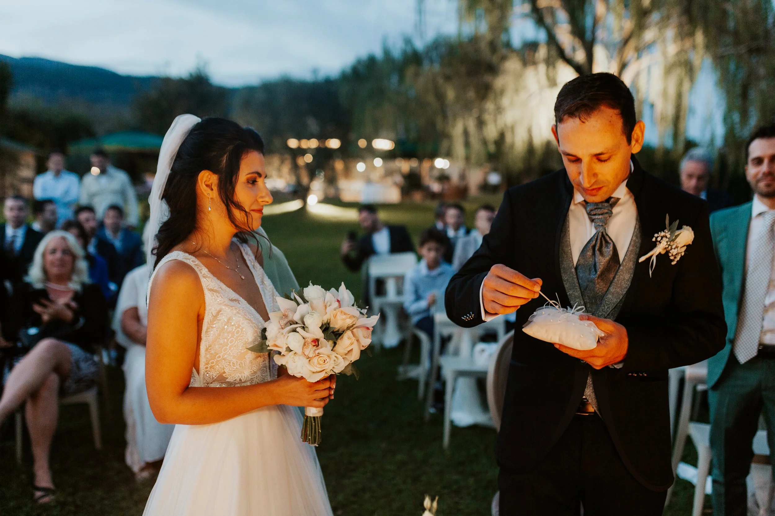 Una coppia di sposi in matrimonio all'aperto con amici e famiglia sullo sfondo, durante il tramonto.
