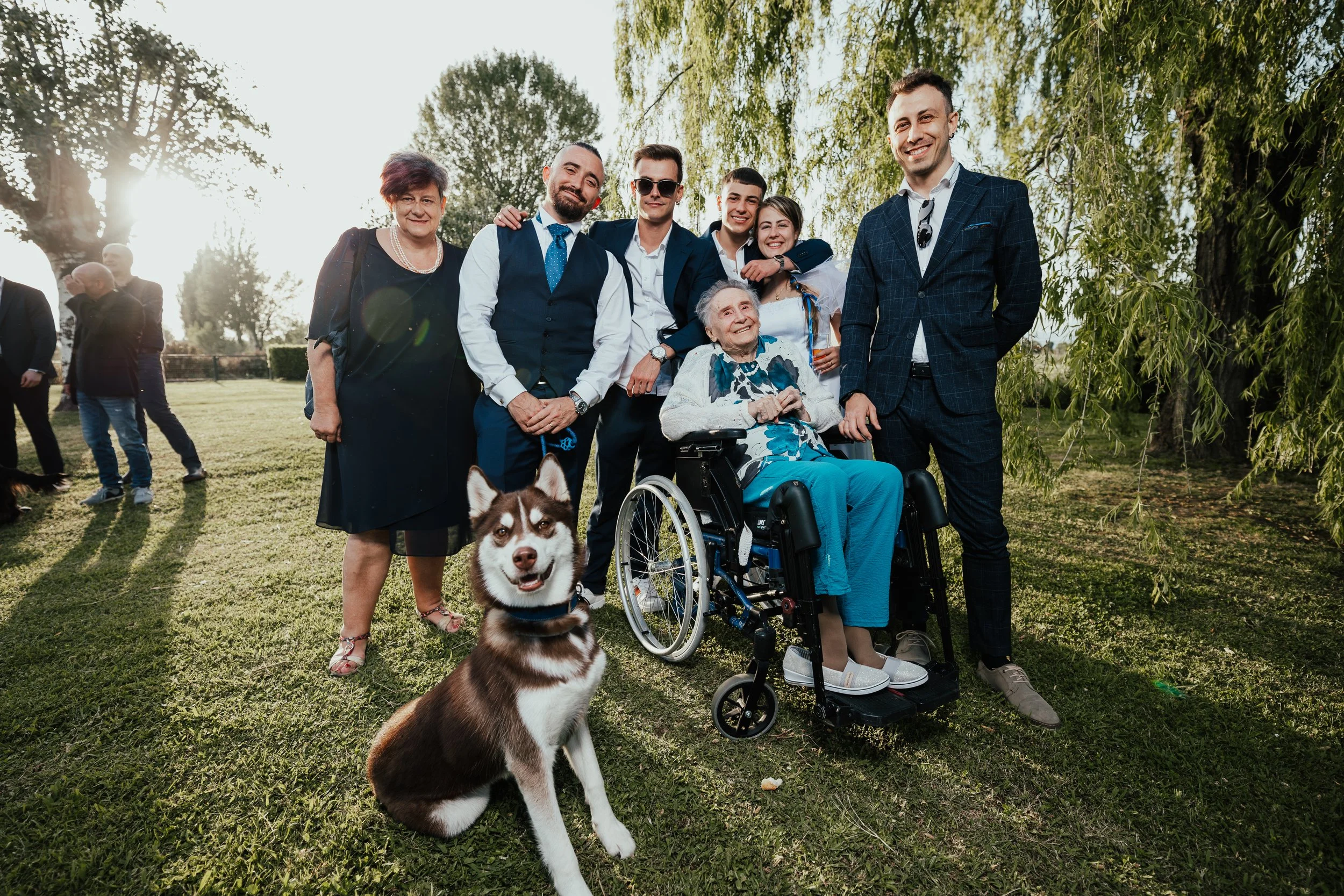 Gruppo di persone sorridenti in un parco con una donna anziana in sedia a rotelle e un cane husky