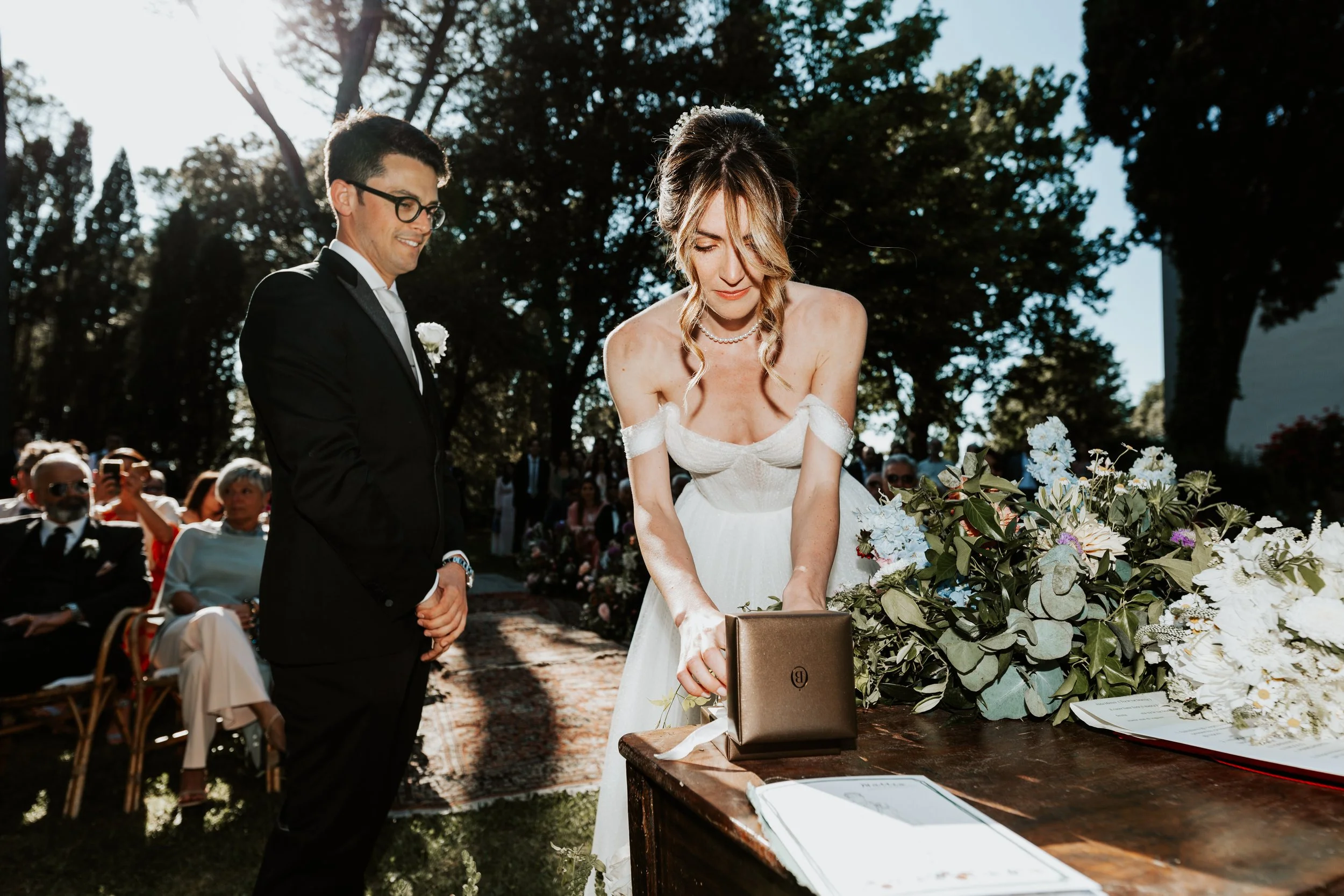 Una coppia di sposi durante il matrimonio all'aperto, la donna firma un documento su un tavolo decorato con fiori, mentre l'uomo la guarda sorridendo, circondati da amici e parenti seduti e in piedi.