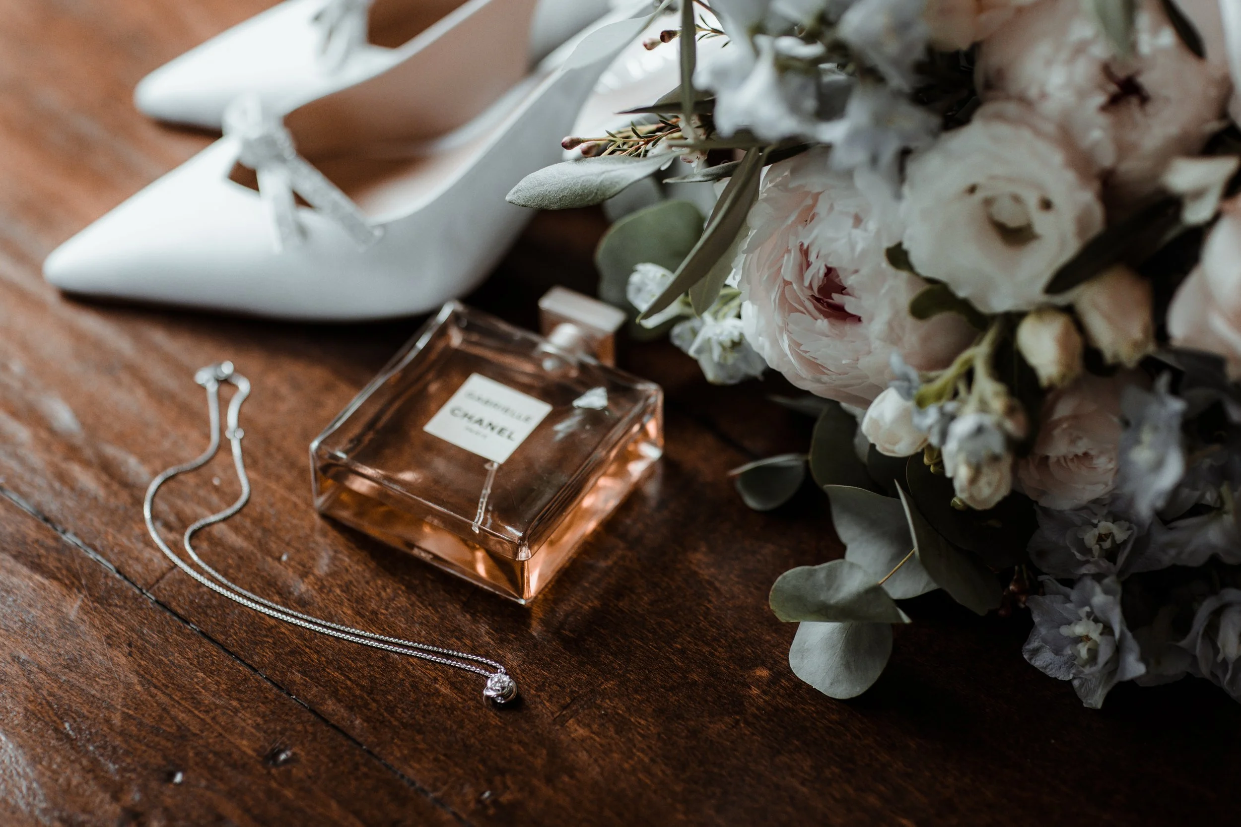 Scarpe bianche col tacco, collana con pendente, boccetta di profumo Chanel e un bouquet di fiori bianchi e verdi su un tavolo di legno.
