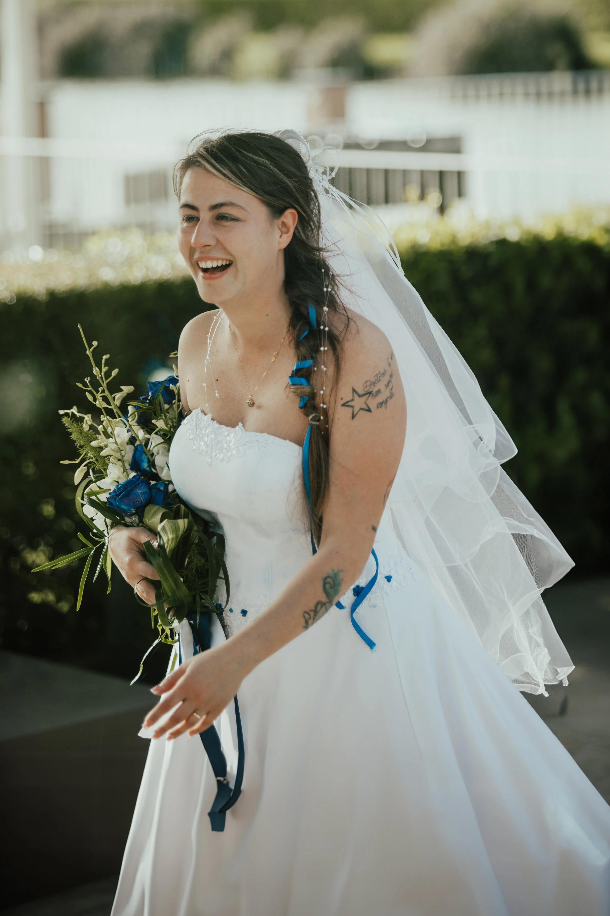 Una sposa sorridente con un abito bianco, un velo e un bouquet di fiori tra cui spiccano fiori blu, all'aperto con sfondo di cespugli e case.