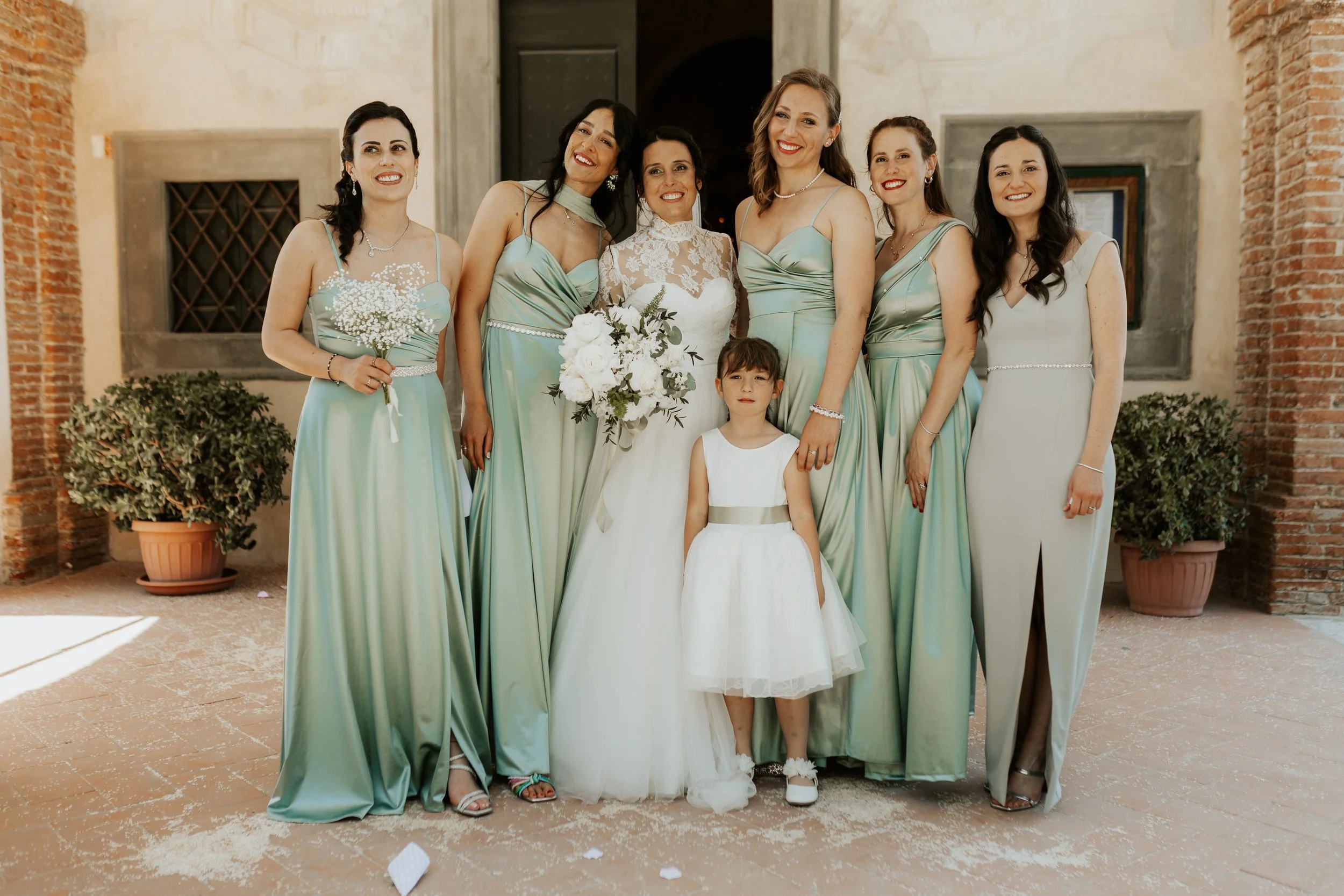 Gruppo di donne in abiti da sera e una bambina in abito bianco, probabilmente a una festa o un matrimonio, con un edificio in stile antico sullo sfondo.