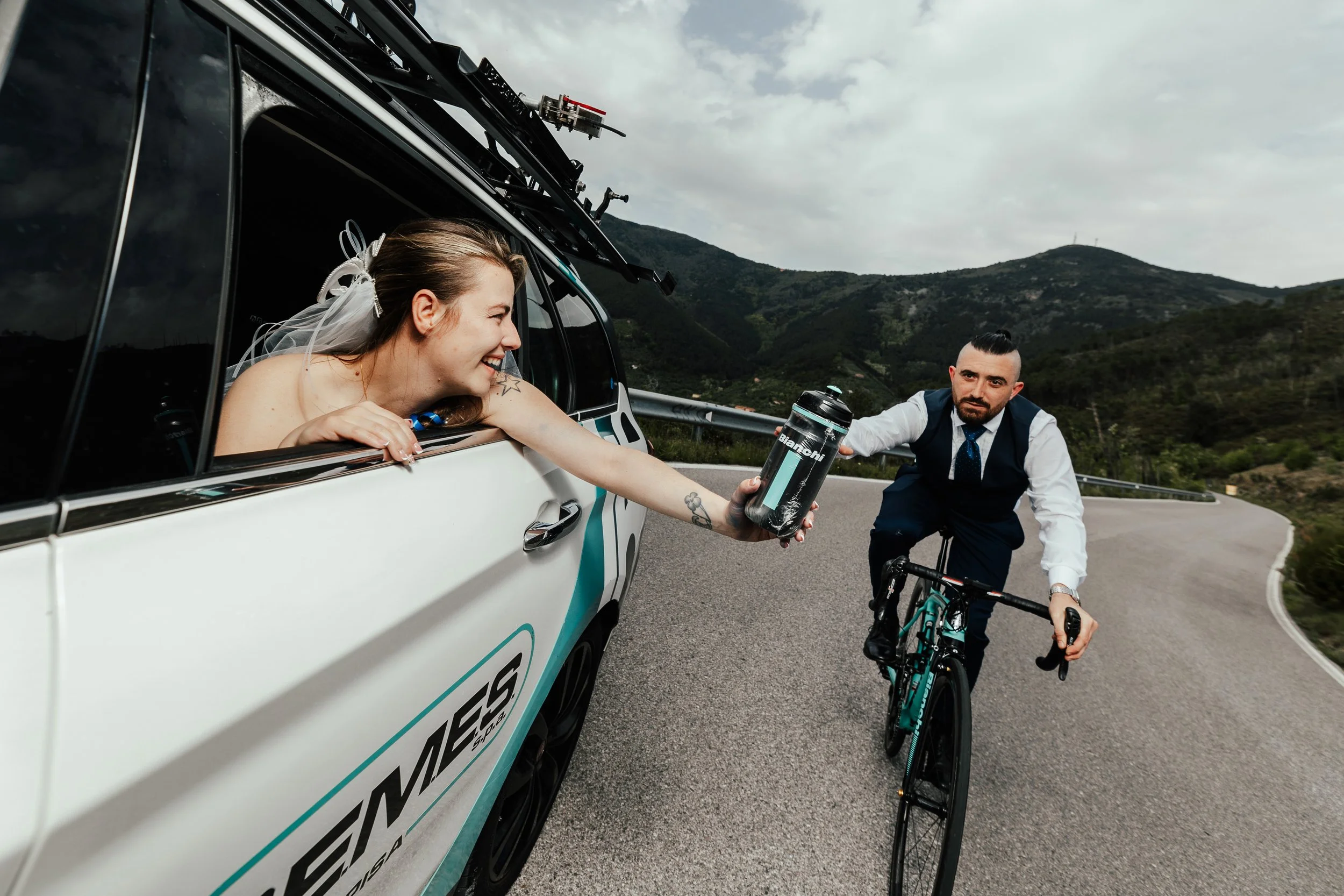 Una donna sorridente in un'auto di produzione racing si passa una borraccia a un uomo in abiti formali che pedala in bicicletta su una curva di montagna.