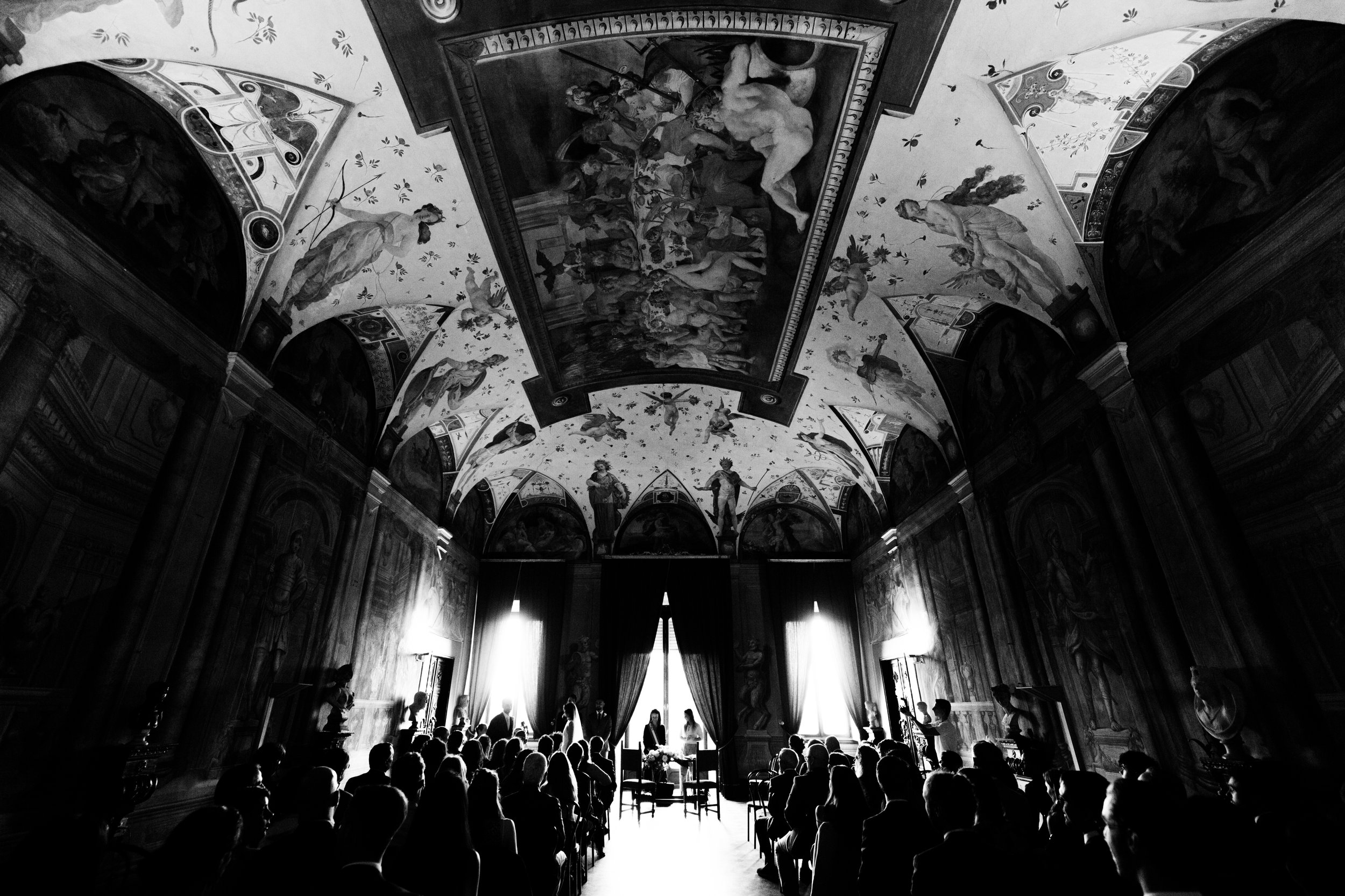 Interno di una sala storica con soffitto affrescato; persone sedute in un evento, luminose finestre sul fondo, grandi tende scure.