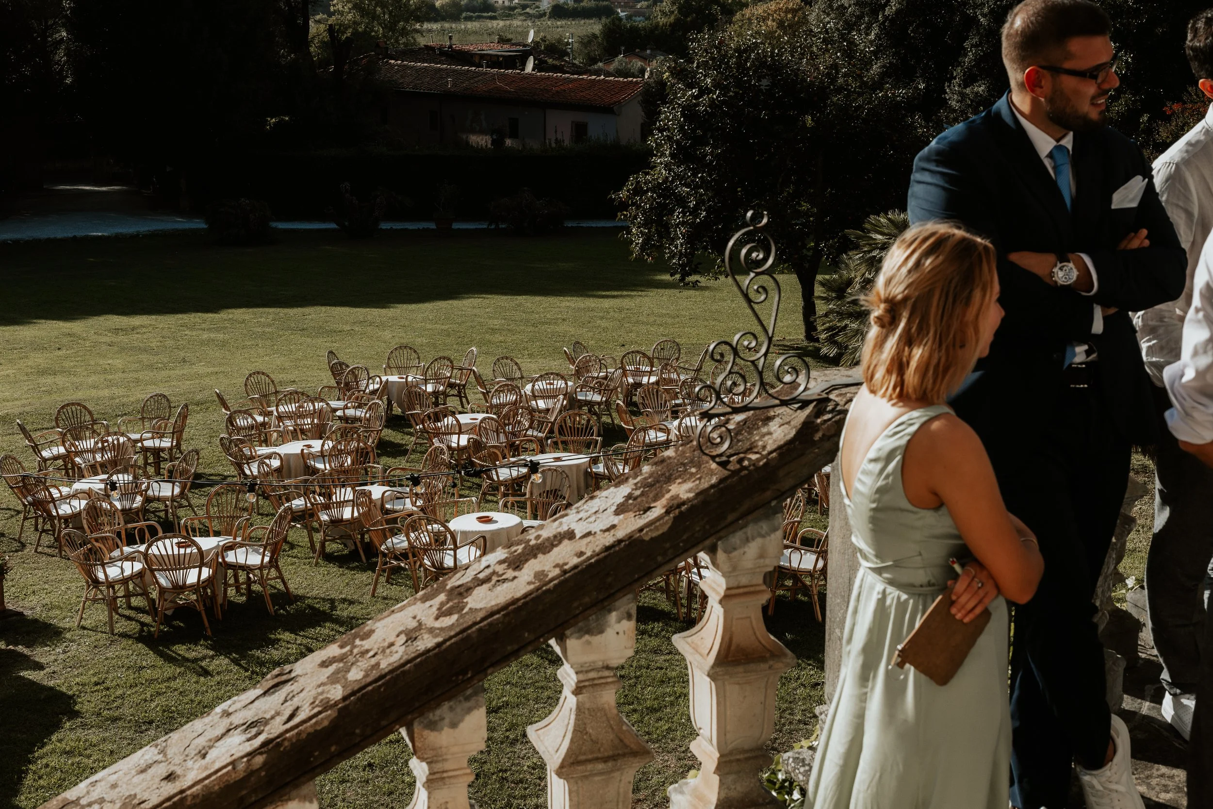 Vista di un giardino con tavoli e sedie di legno pronte per un evento, vista da una terrazza con alcune persone in abiti eleganti. Villa di Corliano