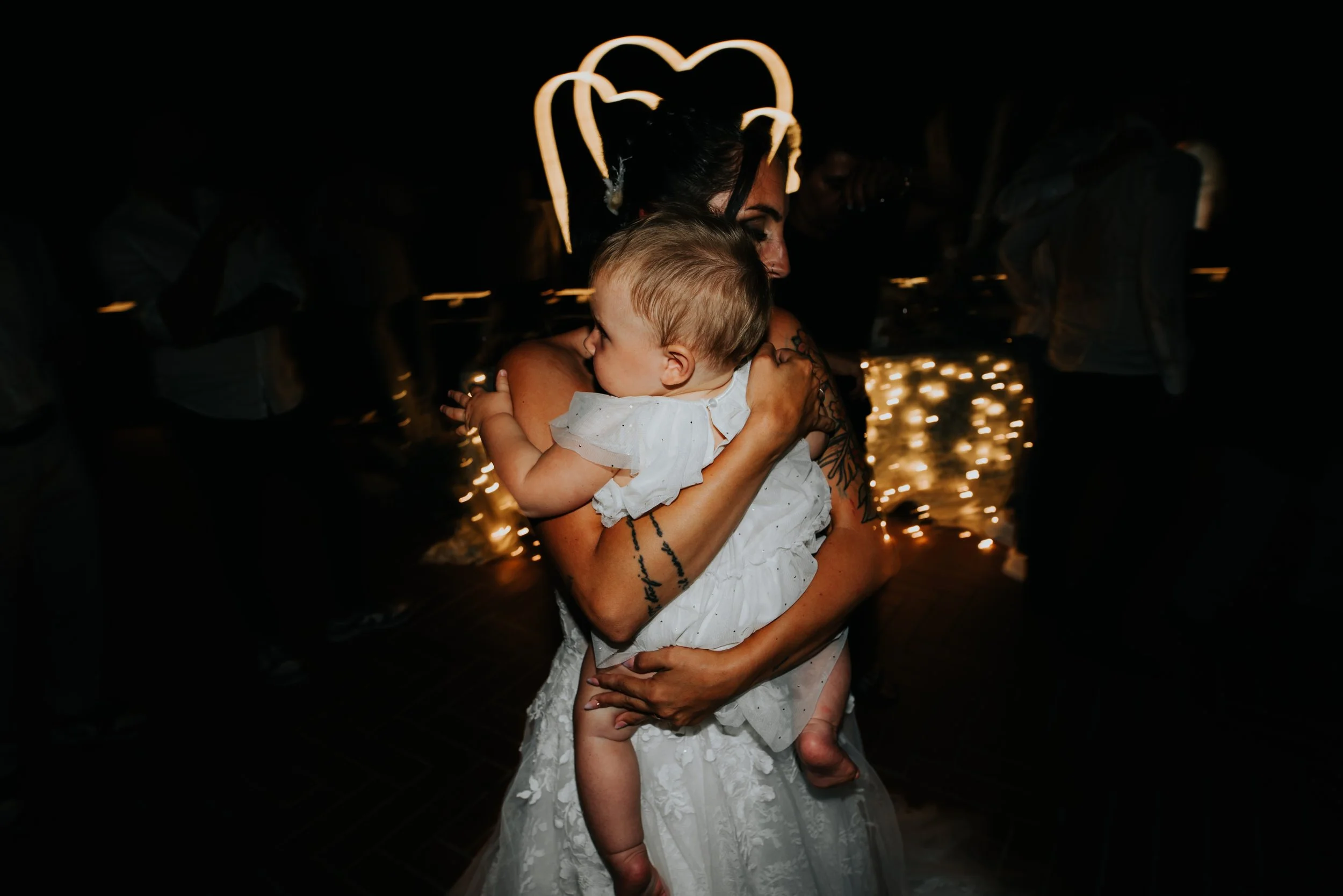 Una donna con un cappello a forma di cuore abbraccia un bambino durante una festa, illuminata da luci natalizie, in un ambiente oscuro.