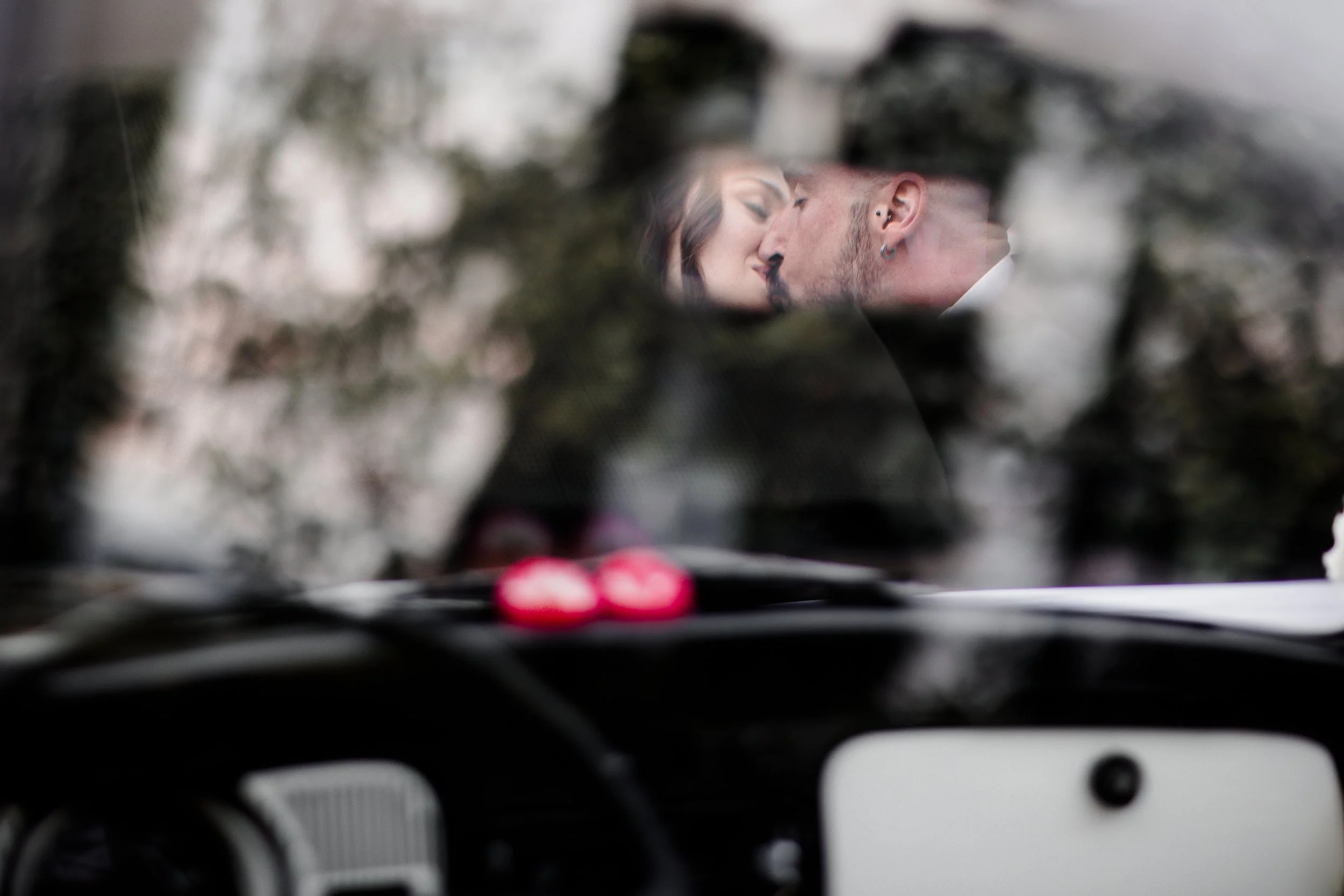 Riflesso di una coppia che si bacia, visto attraverso uno specchietto retrovisore di un'auto, con alcuni oggetti rossi sul cruscotto.