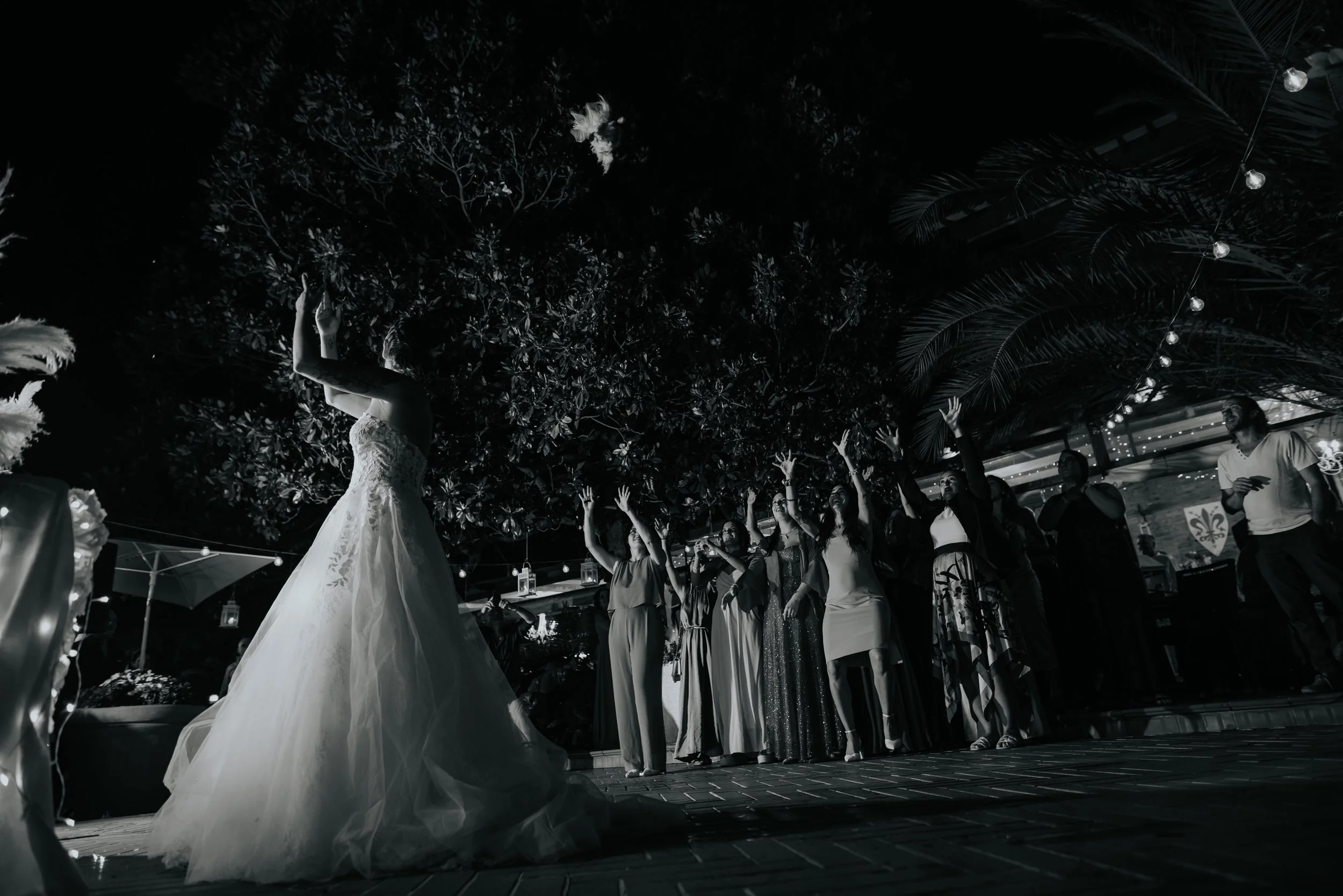 Donna in abito da sposa che lancia il bouquet a un gruppo di donne sorridenti durante una cerimonia di matrimonio all'aperto di notte, con luci decorative e vegetazione sullo sfondo.