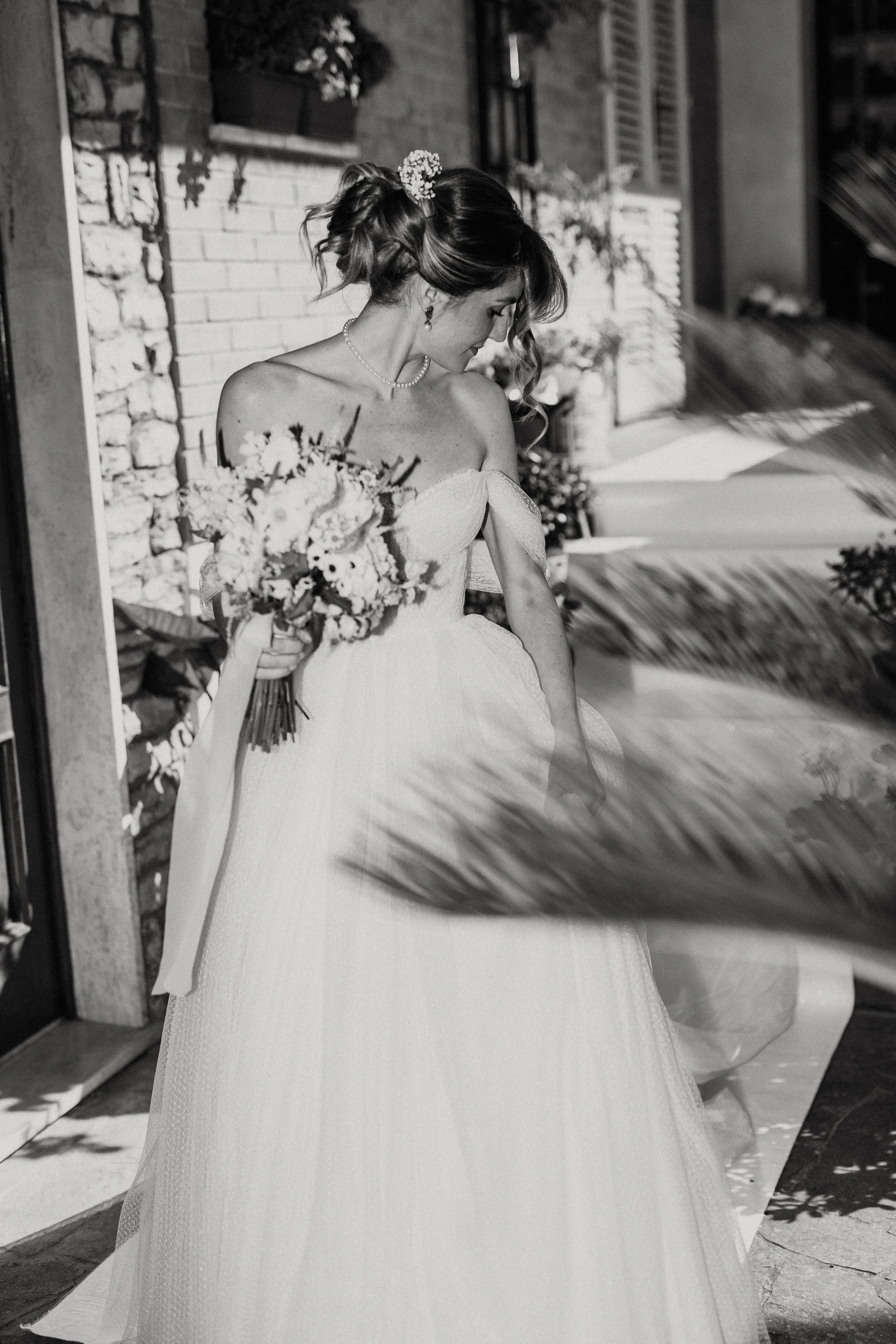 Donna in abito da sposa con bouquet di fiori, che si trova all'aperto vicino a un muro di mattoni, con piante e fiori sullo sfondo.