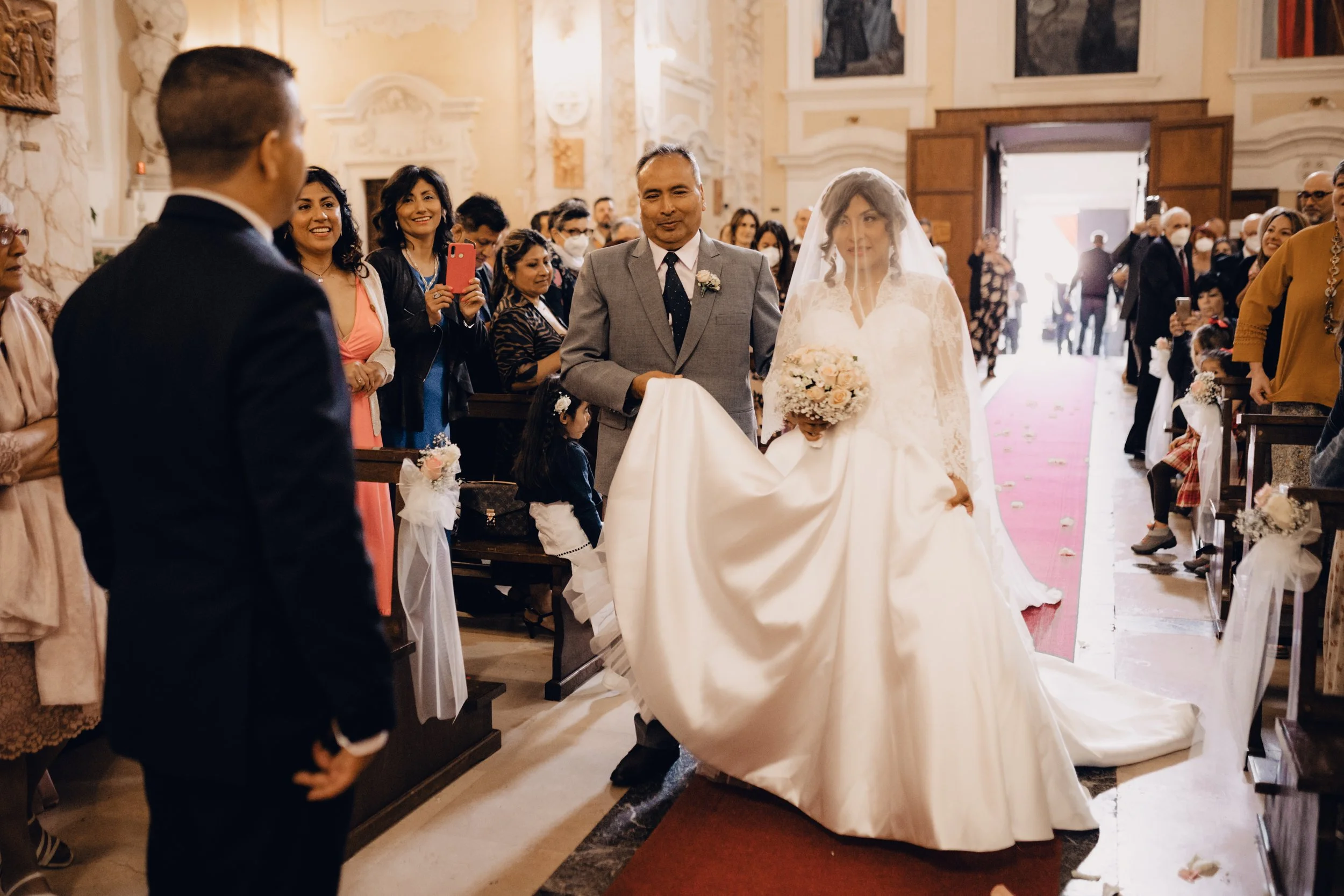 Una sposa cammina nell'altare con il suo padre in chiesa, circondata da amici e famiglia che la fotografano e la guardano con gioia.