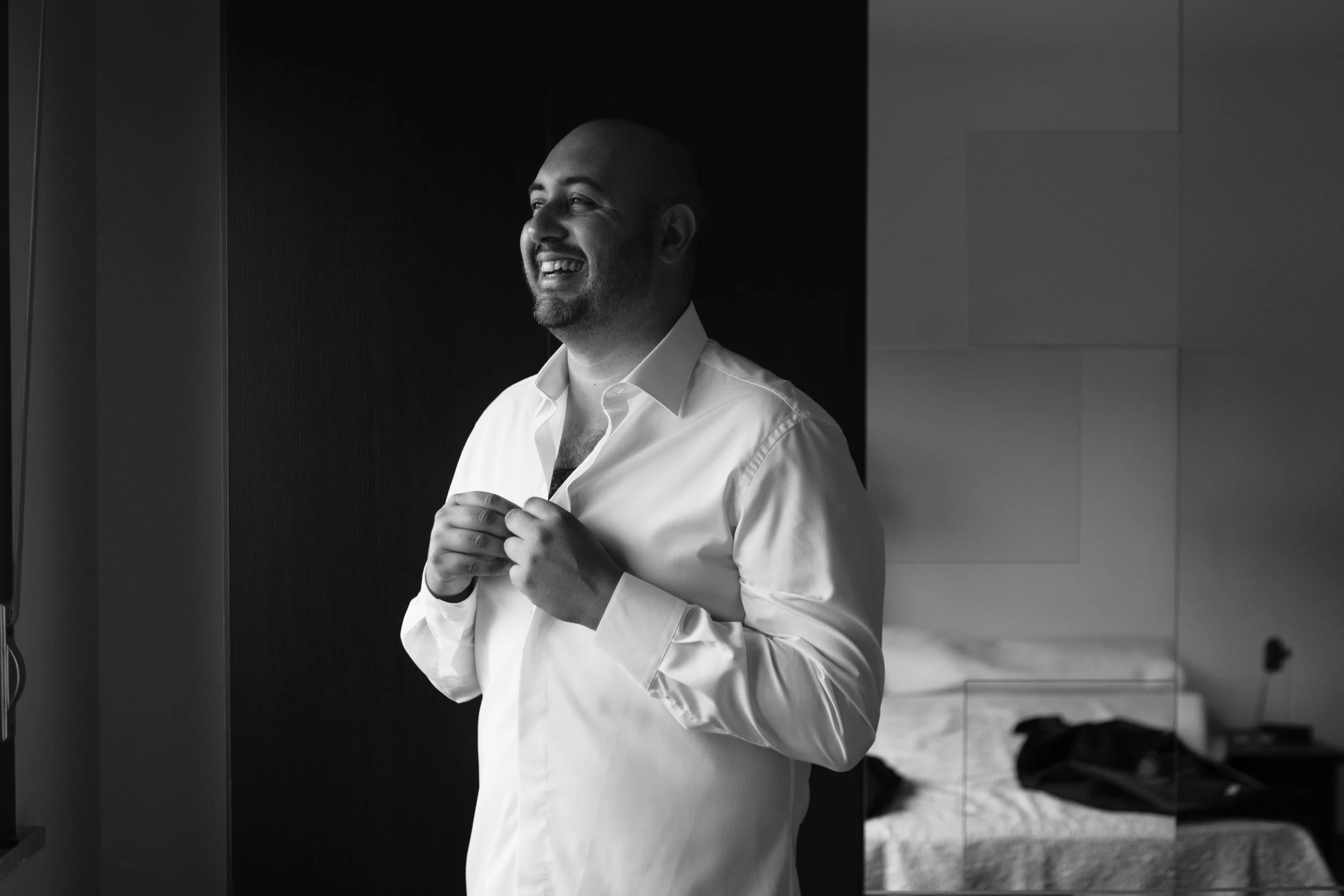 Uomo che si veste con camicia bianca in una stanza moderna con pareti chiuse e letto sullo sfondo.