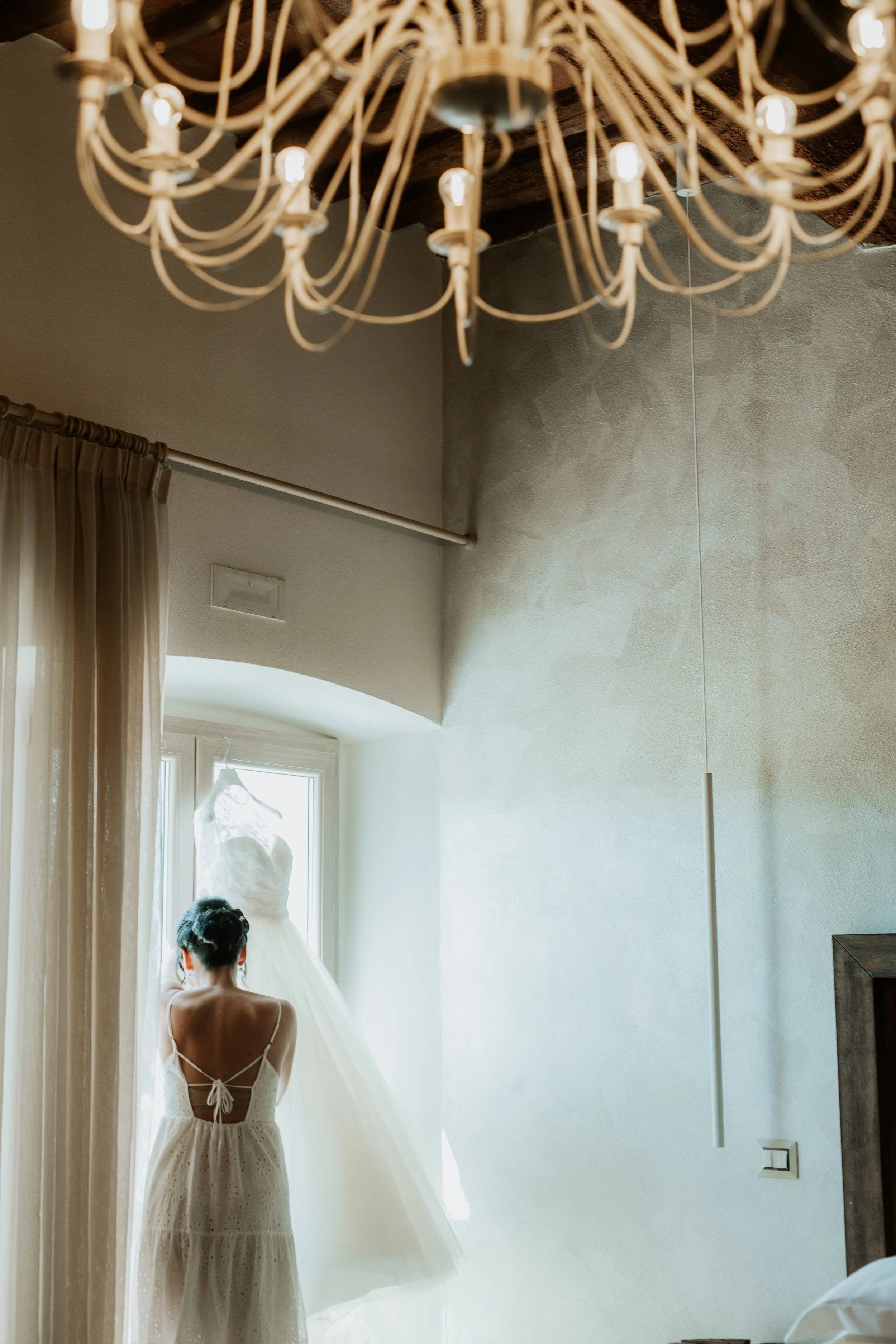 Una donna vestita con un abito bianco guarda una gonna da sposa appesa alla finestra in una stanza luminosa.