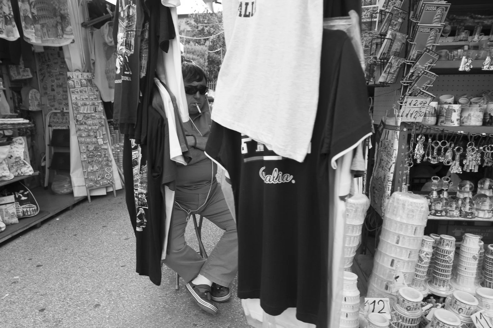 Uomo con occhiali da sole seduto tra bancarelle di oggetti di souvenirs e abbigliamento, in un mercato all'aperto.