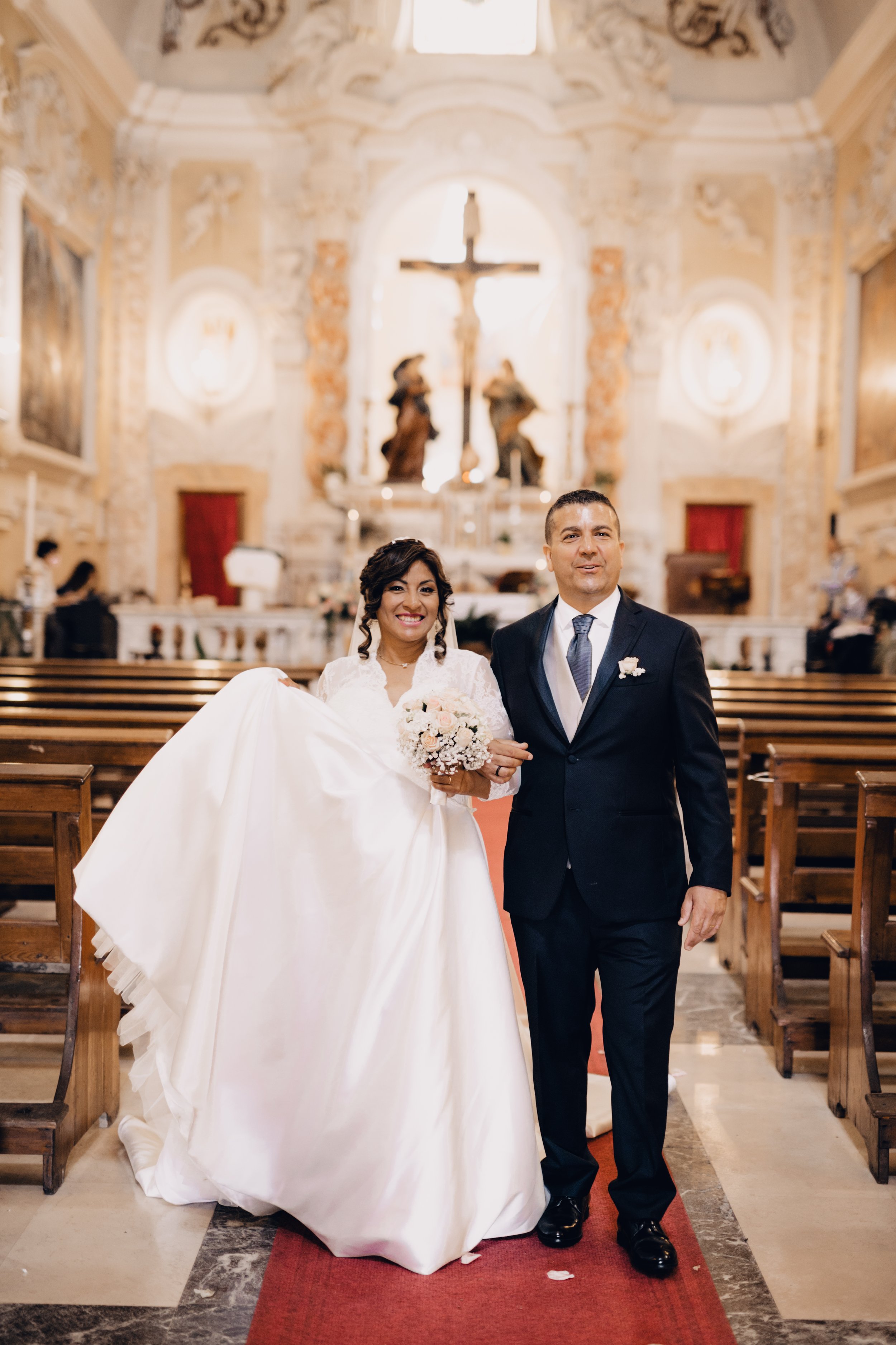 Sposi in chiesa con l'altare sullo sfondo, donna con abito da sposa bianco che tiene un bouquet di fiori, uomo in tuxedo nero, entrambi sorridenti.