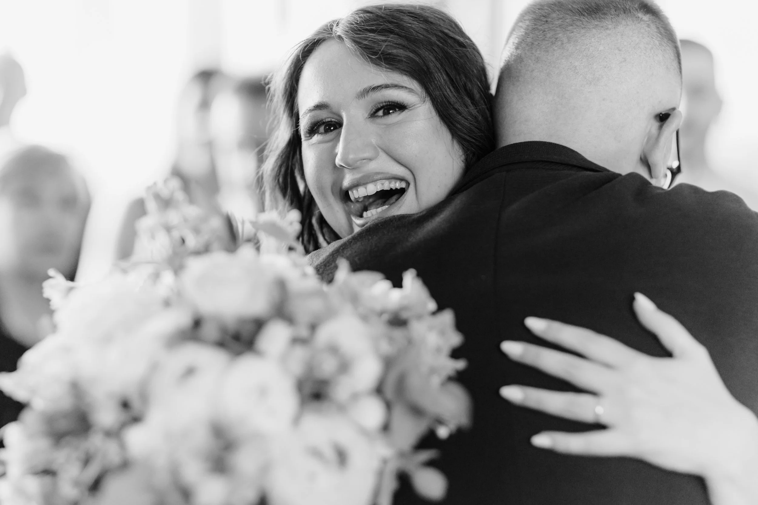 Donna e uomo che si abbracciano, sorridendo, con bouquet di fiori in primo piano, in un contesto cerimoniale, in bianco e nero.