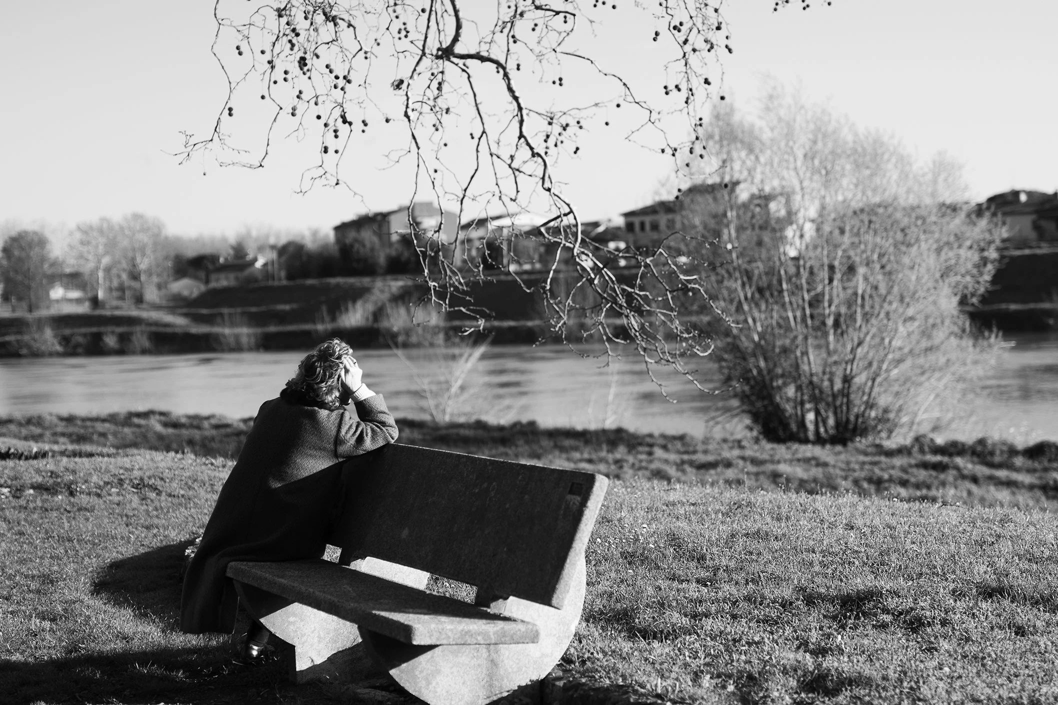 Una donna seduta su una panchina in un parco, in un paesaggio naturale con un fiume, alberi spogli e case sullo sfondo, in bianco e nero.