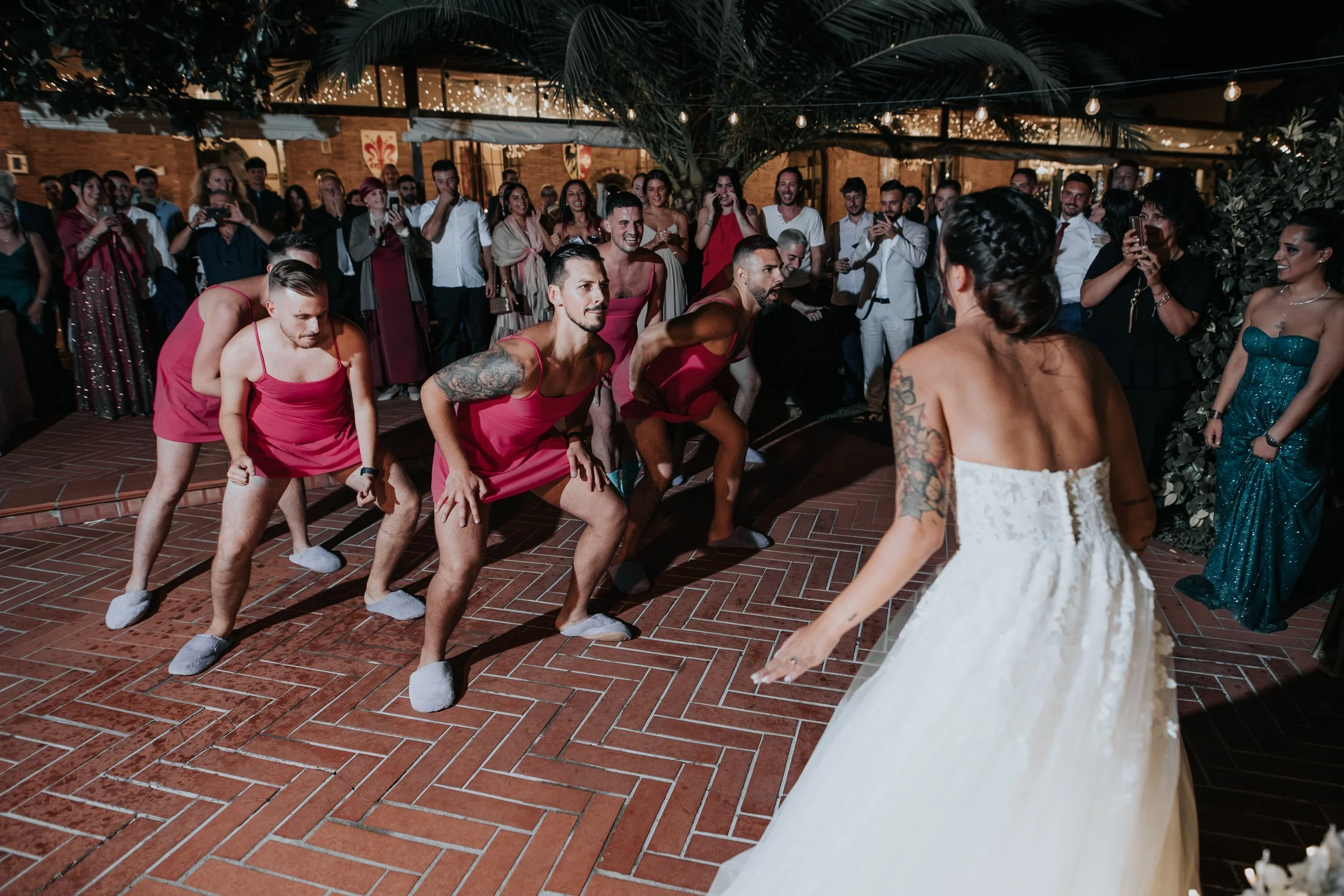 Coppia di sposi che balla di fronte a un gruppo di uomini che indossano abiti rosa e ciabatte, durante una cerimonia di matrimonio all'aperto con decorazioni luminose.