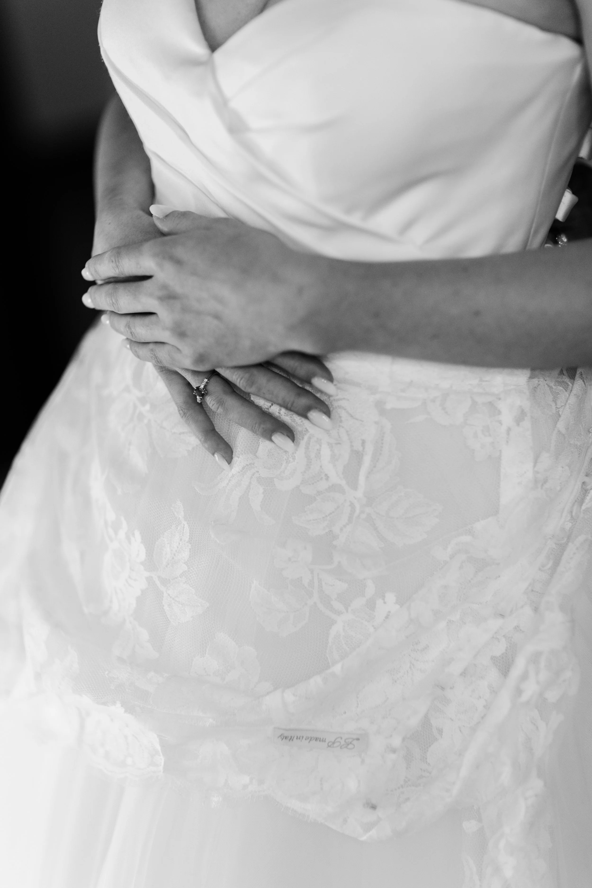 Immagine in bianco e nero di una donna che indossa un abito da sposa, con le mani incrociate sul ventre, una delle quali mostra un anello di matrimonio.