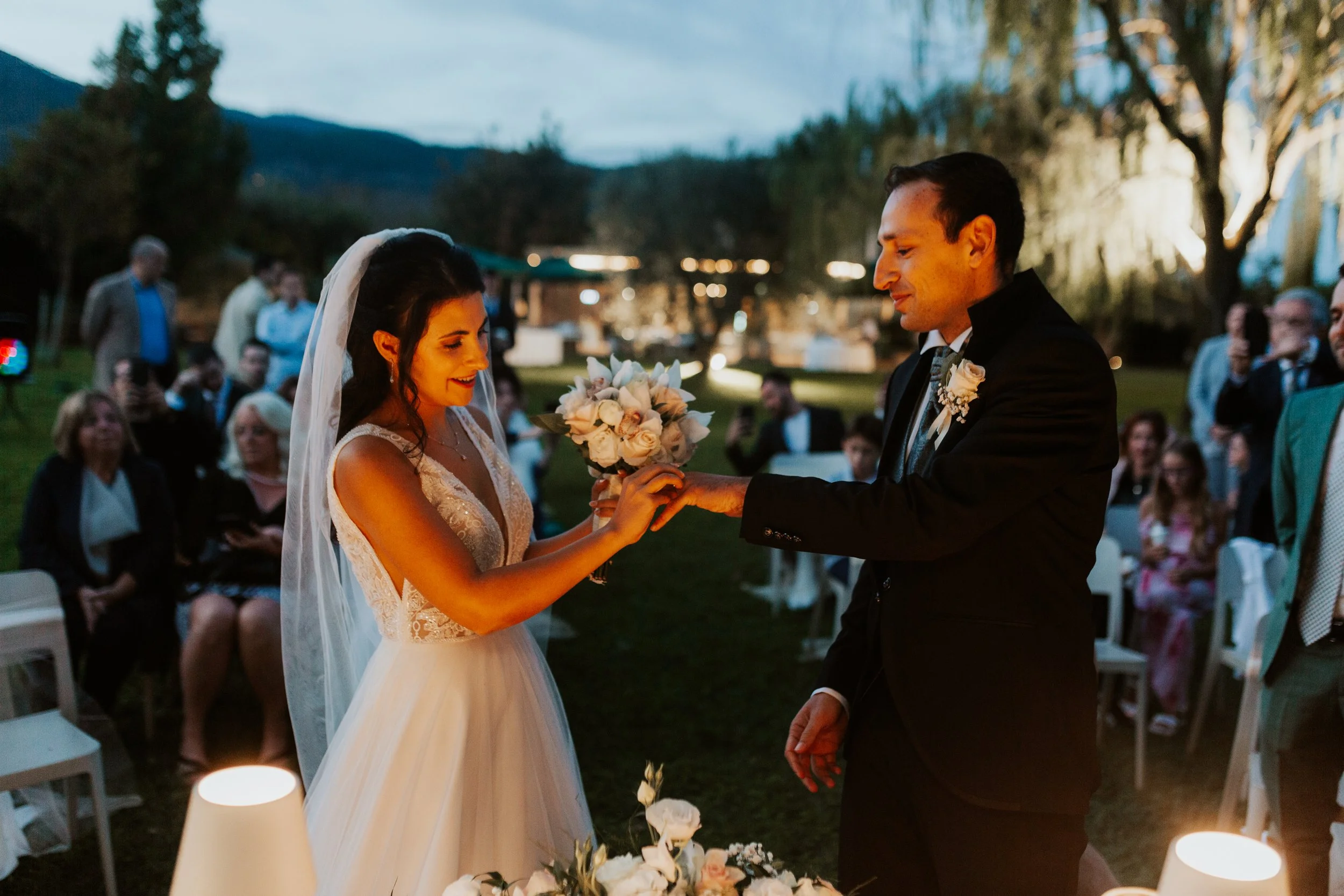 Sposi durante il matrimonio all'aperto in serata, scambio di anelli, con amici e parenti seduti intorno, luci soffuse, atmosfera romantica
