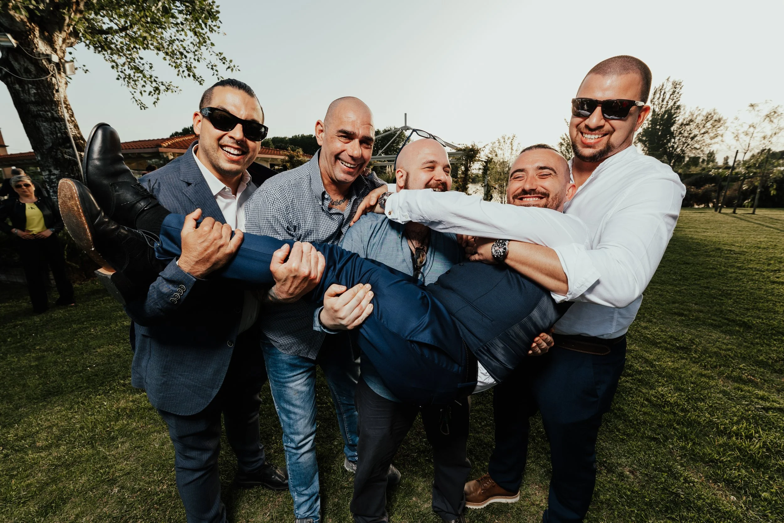 Gruppo di uomini sorridenti che si divertono trascinando un altro uomo a terra in un'area verde all'aperto.