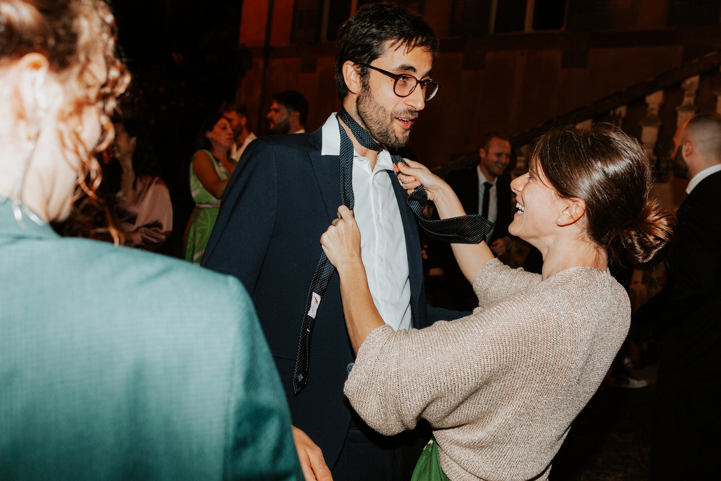 Gruppo di persone che ride e si diverte a una festa, con un uomo in giacca e donna che lo aiuta a indossare una cravatta