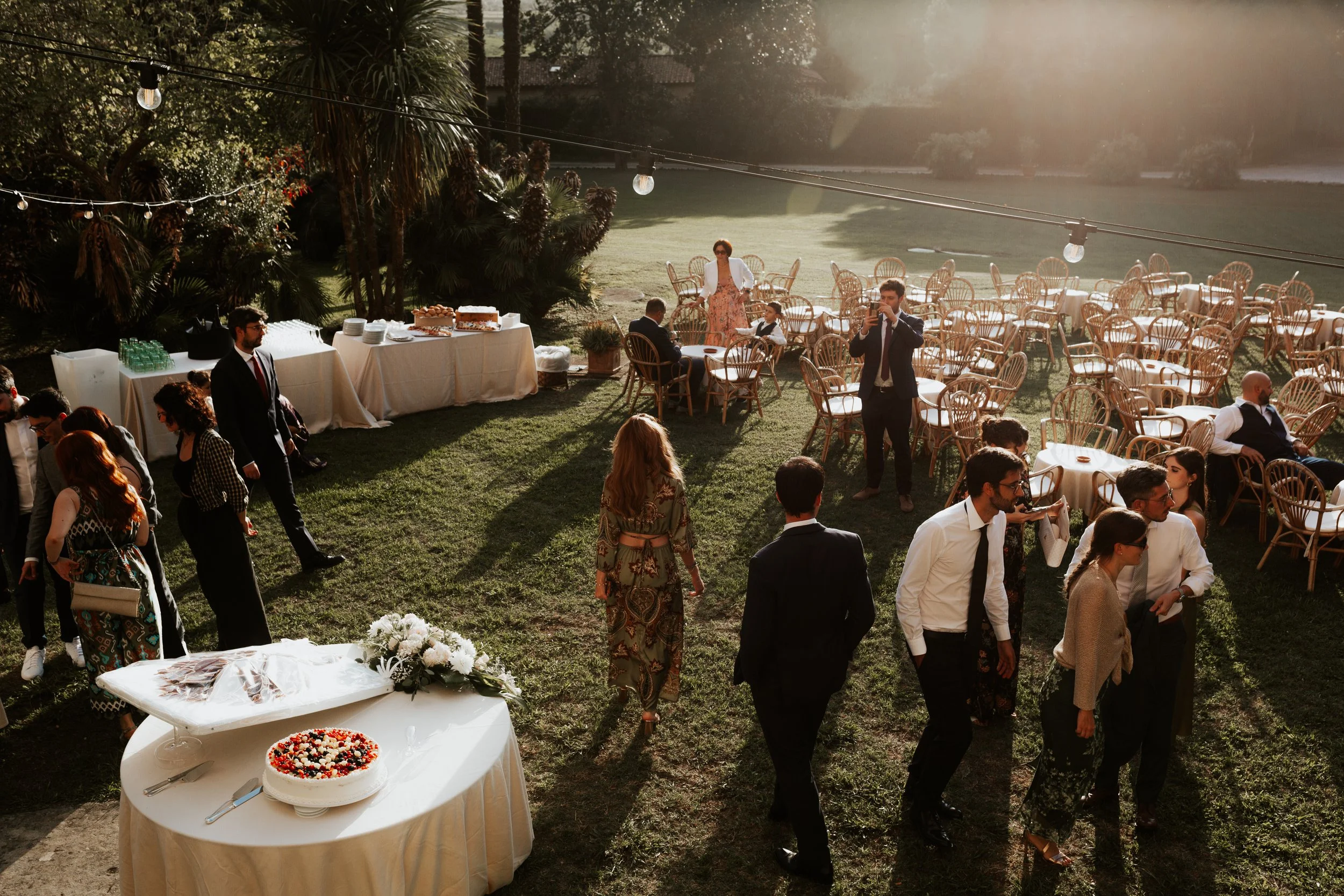 Celebrità e amici che socializzano a un ricevimento all'aperto durante il tramonto, con tavoli, cibo e decorazioni eleganti in un giardino.