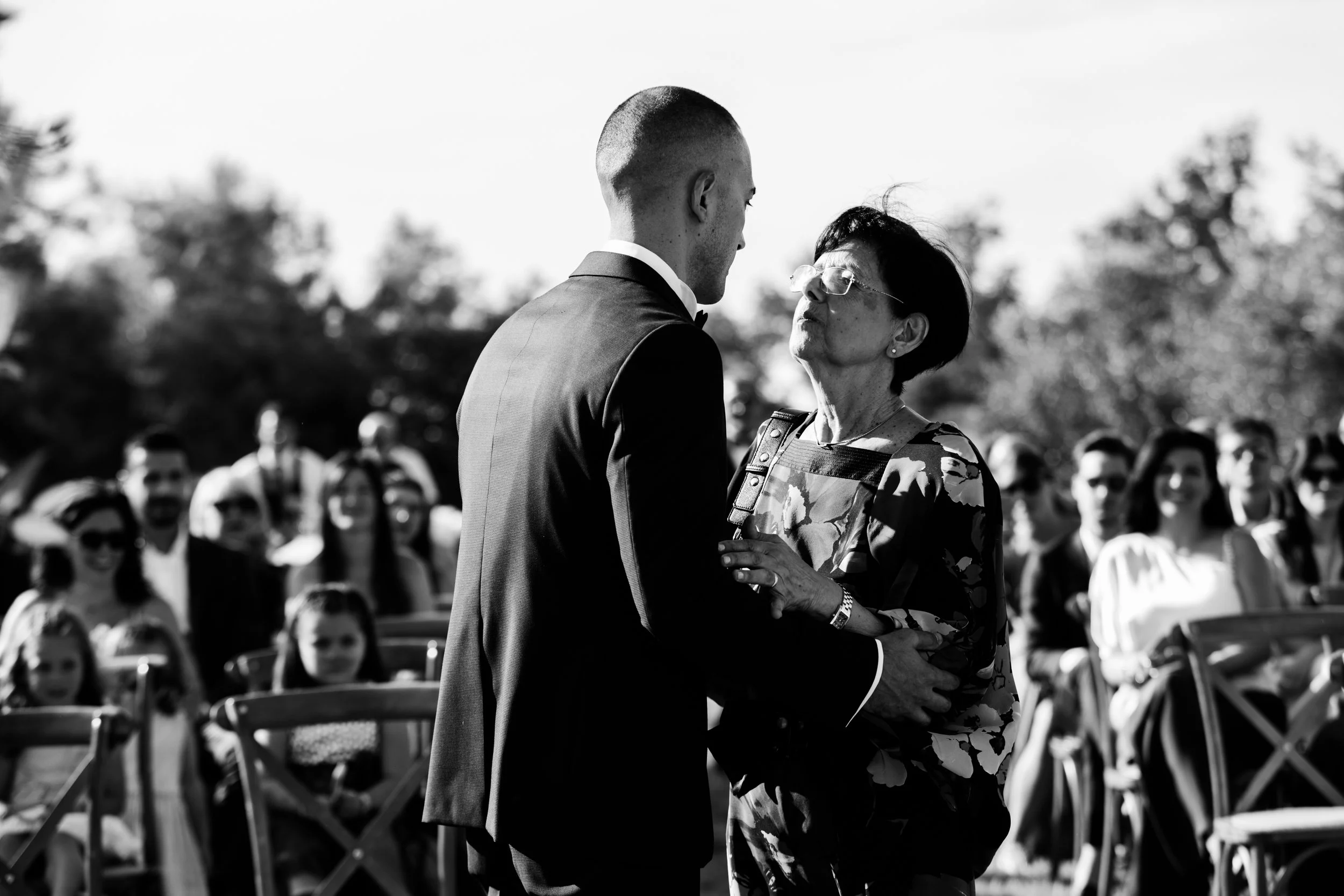 Un uomo elegantemente vestito di nero durante un matrimonio, che tiene per mano una donna anziana con occhiali e abbigliamento floreale, davanti a un gruppo di persone sedute e sorridenti all'aperto.