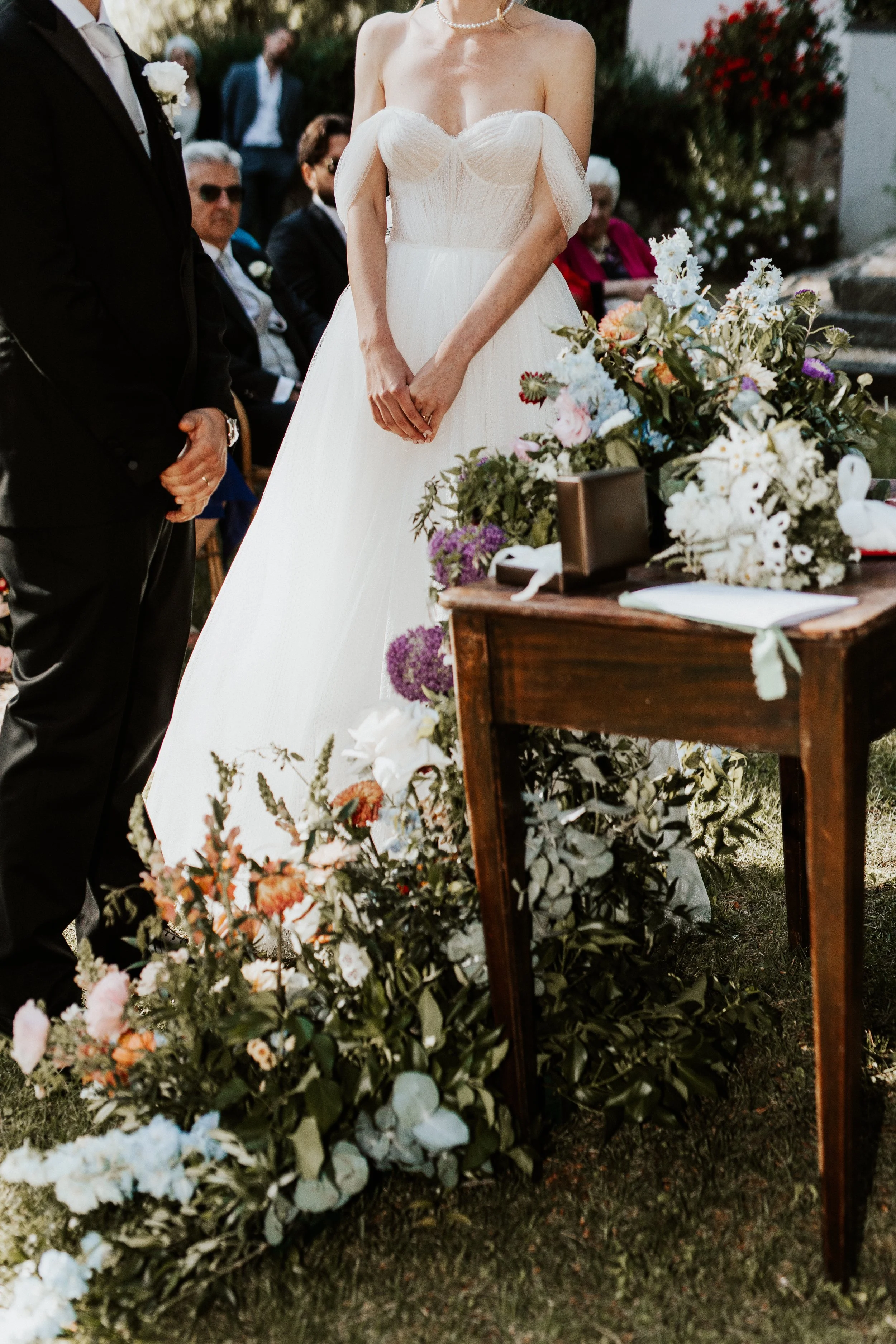 Cerimonia di matrimonio con sposa in abito bianco e sposo in completo nero, circondati da fiori e invitati seduti. Villa dal Pozzo