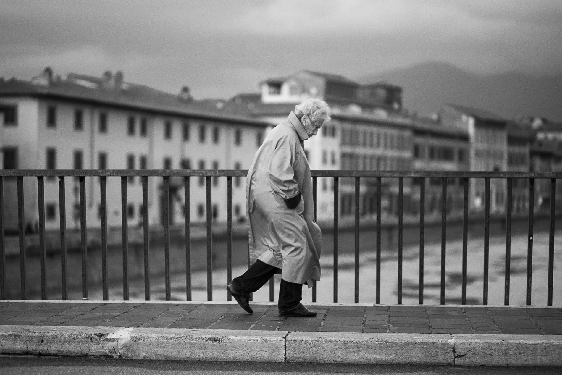Una persona anziana cammina lungo un marciapiede sotto un cielo nuvoloso, in una scena urbana in bianco e nero.