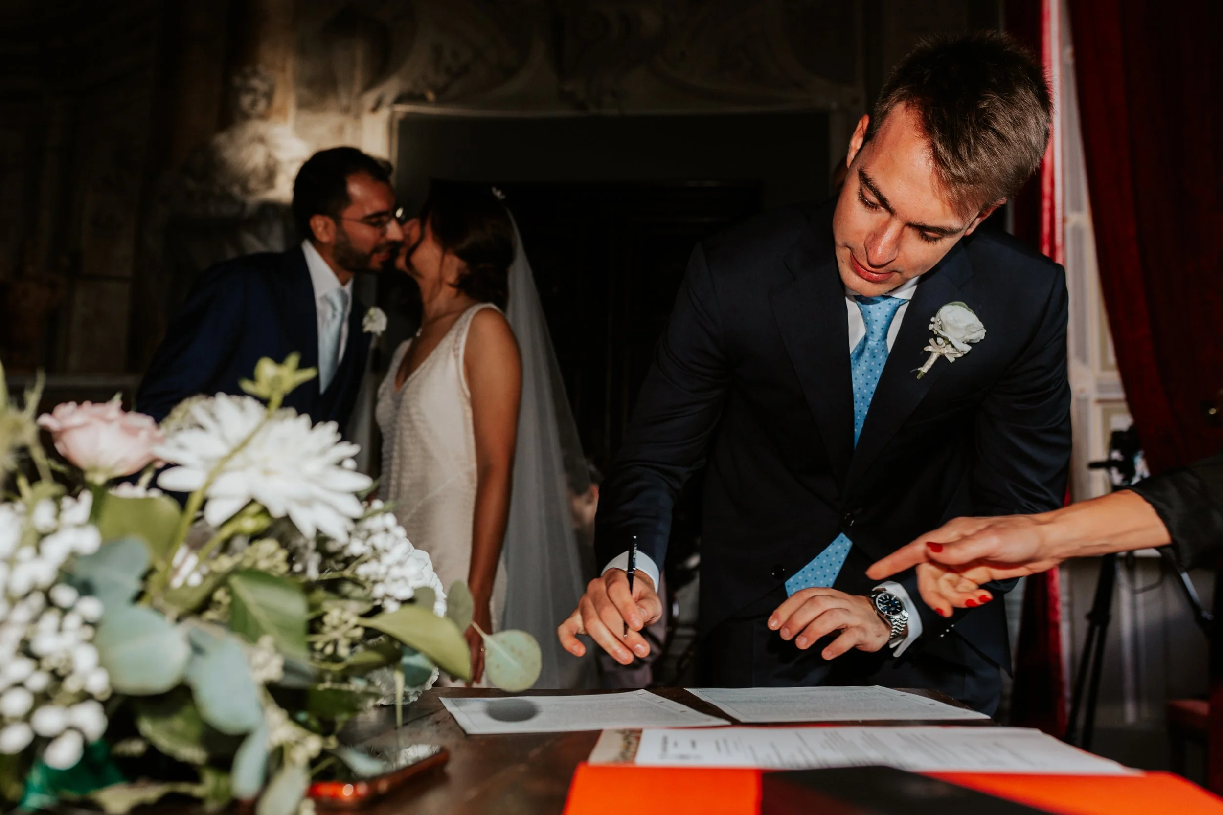 Un uomo firma un documento durante un matrimonio, con un bouquet di fiori in primo piano e una coppia di sposi sullo sfondo che si bacia.