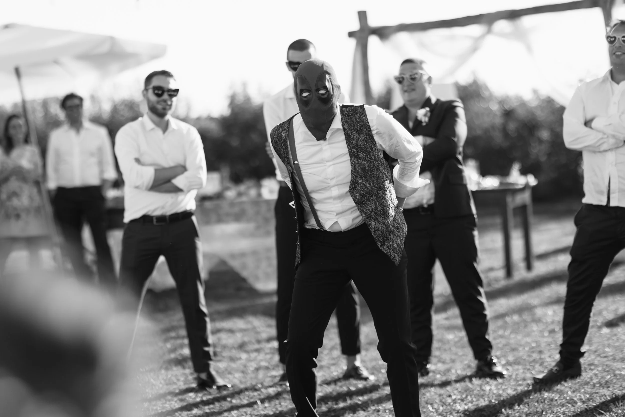 Un uomo con maschera di Deadpool, abbigliamento elegante, balla durante un evento all'aperto con altre persone che indossano abiti formali, in bianco e nero.