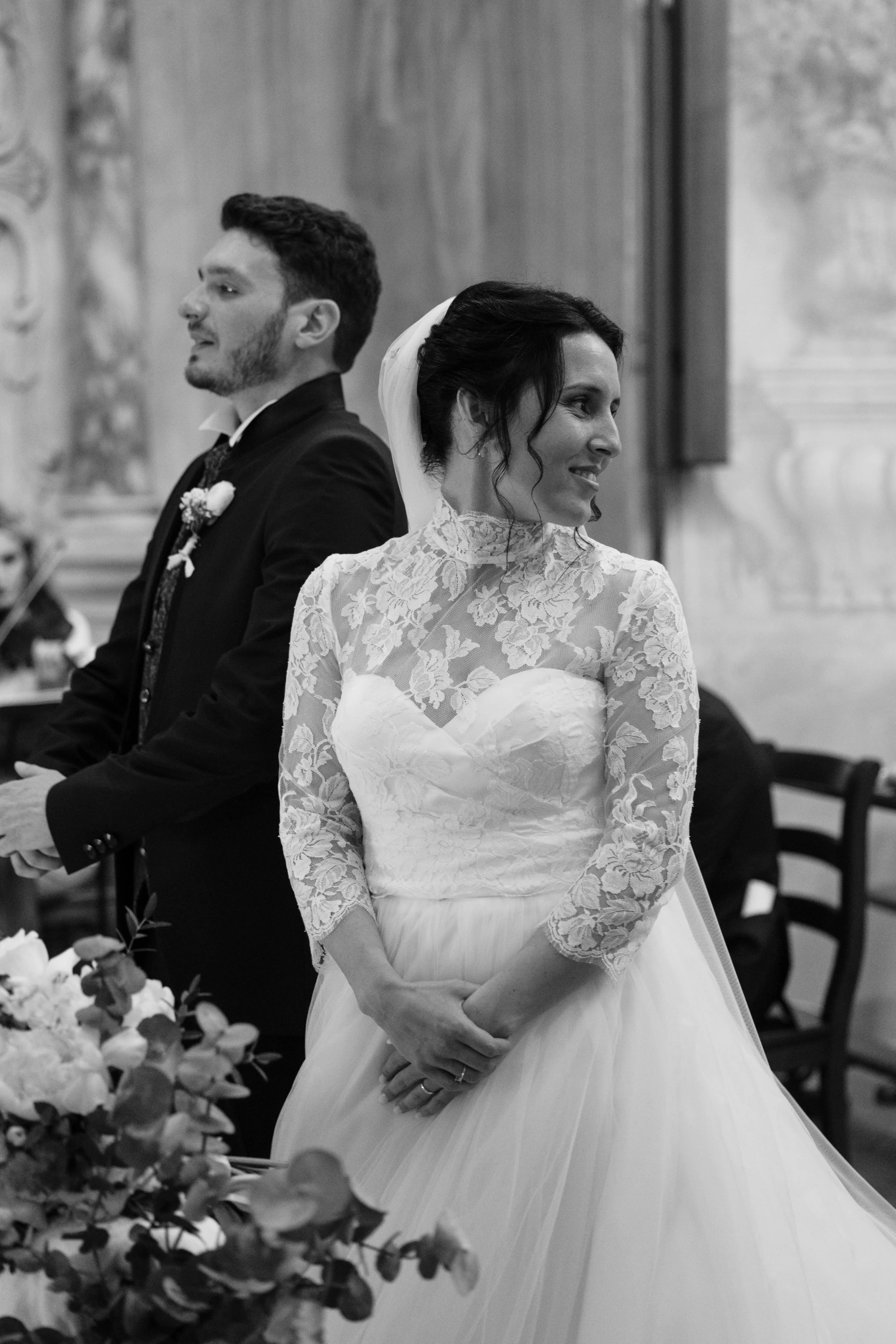 Una coppia di sposi durante il matrimonio, la donna indossa un abito bianco con top in pizzo e la gonna ampia, l'uomo indossa un abito scuro. Sono in chiesa, in piedi, con sfondo di arredi sacri e altre persone.