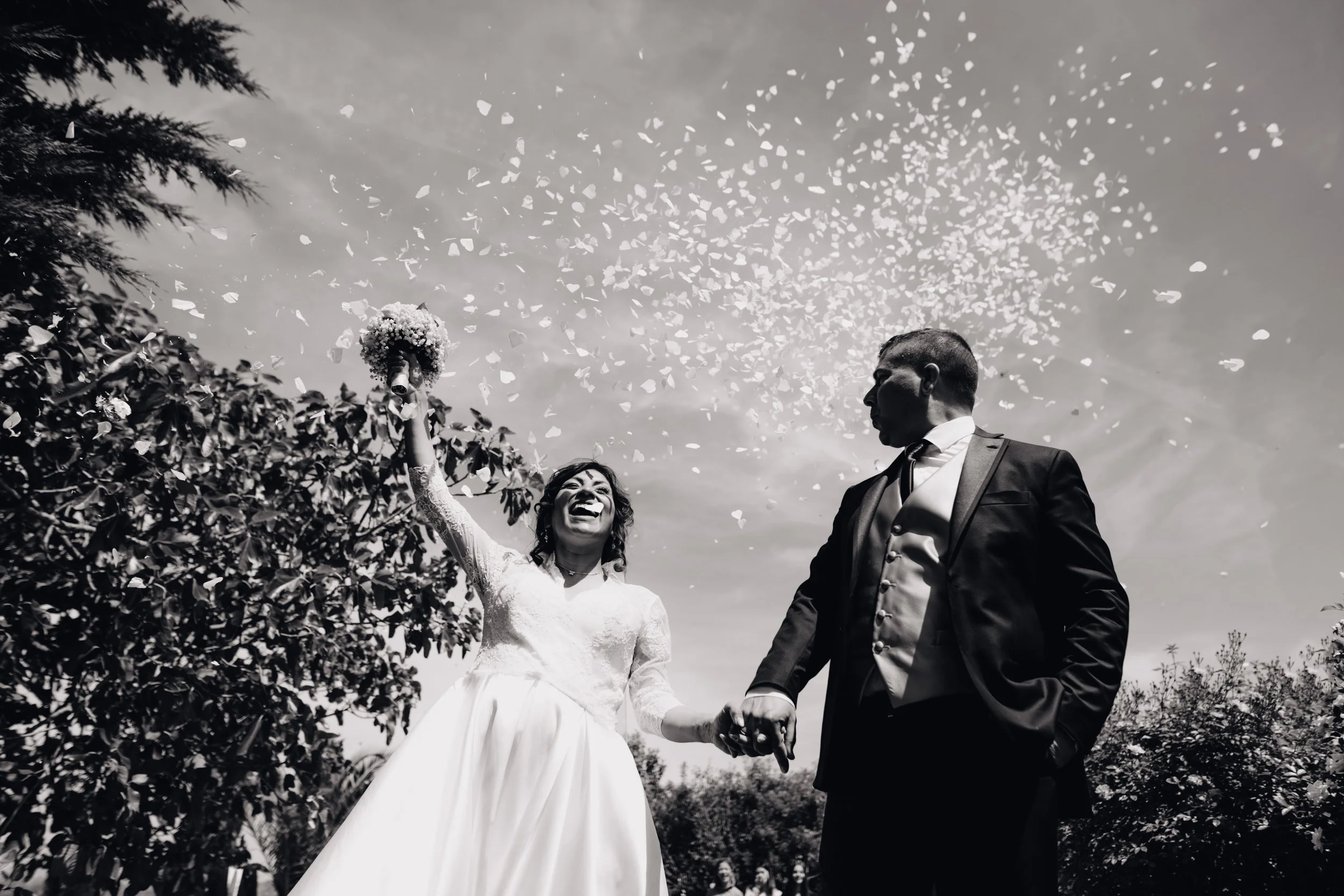 Una coppia di sposi felice si tiene per mano sotto un cielo sereno, con petali che cadono, durante il matrimonio.