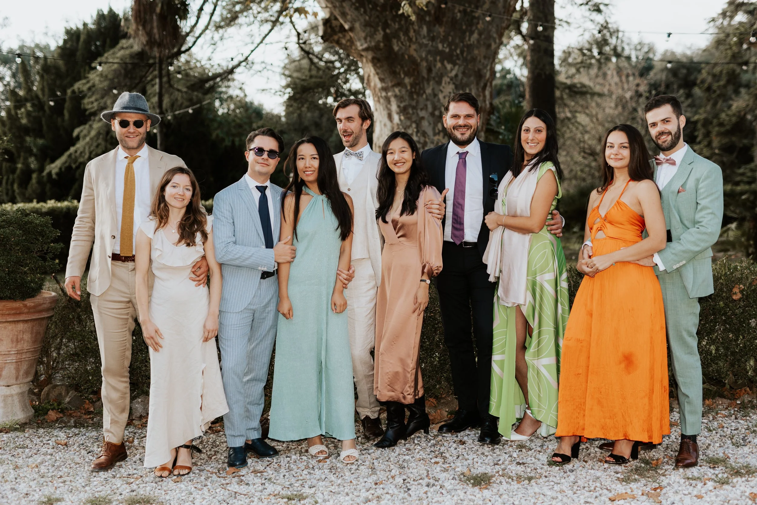Un gruppo di persone in abiti eleganti si trova all'aperto, sorridendo, sotto un grande albero, probabilmente a una festa o un matrimonio.