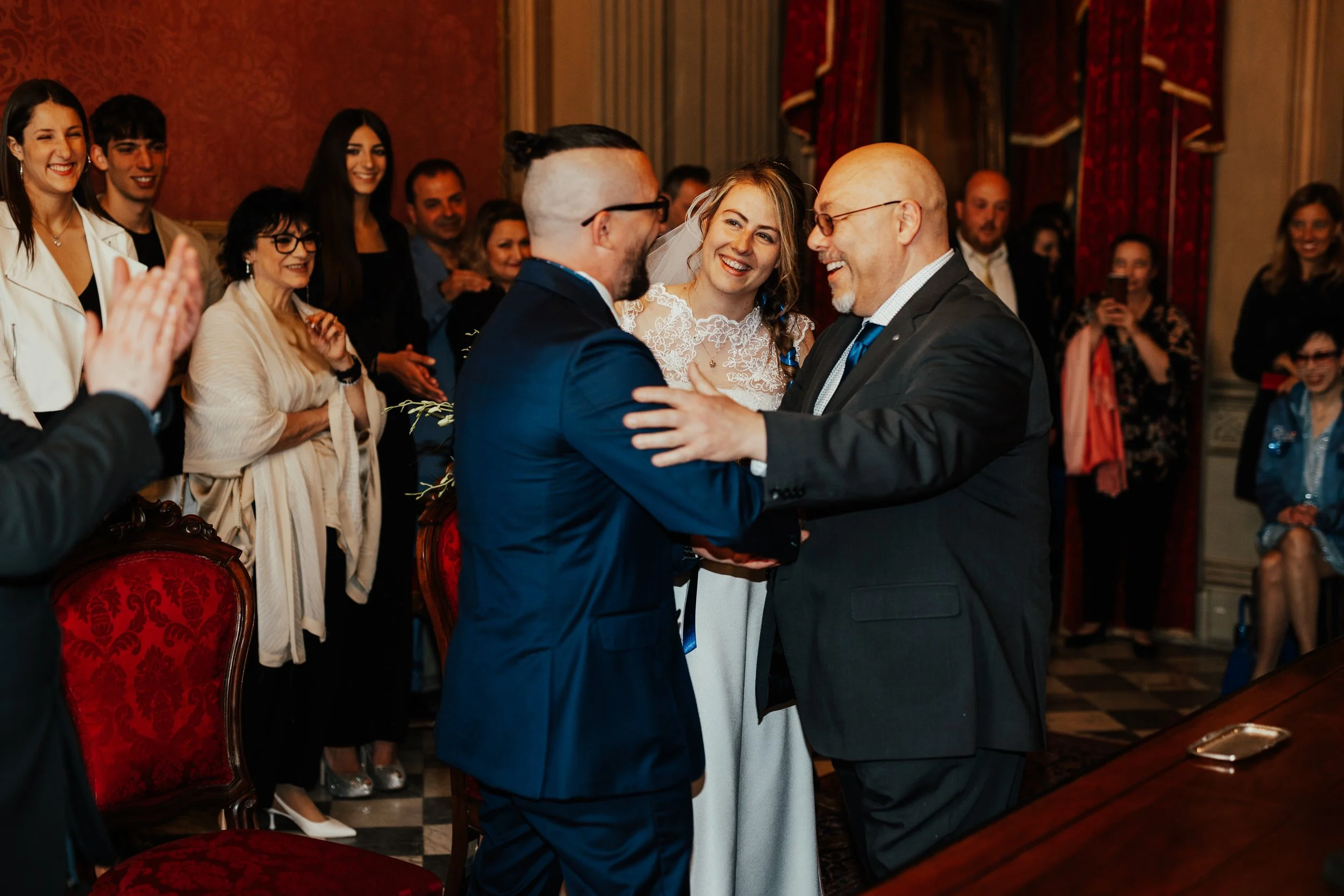 Cerimonia di matrimonio con sposo, sposa e famiglia che si abbracciano, mentre gli invitati sorridono e guardano, ambiente elegante con arredamenti classici e drappi rossi.