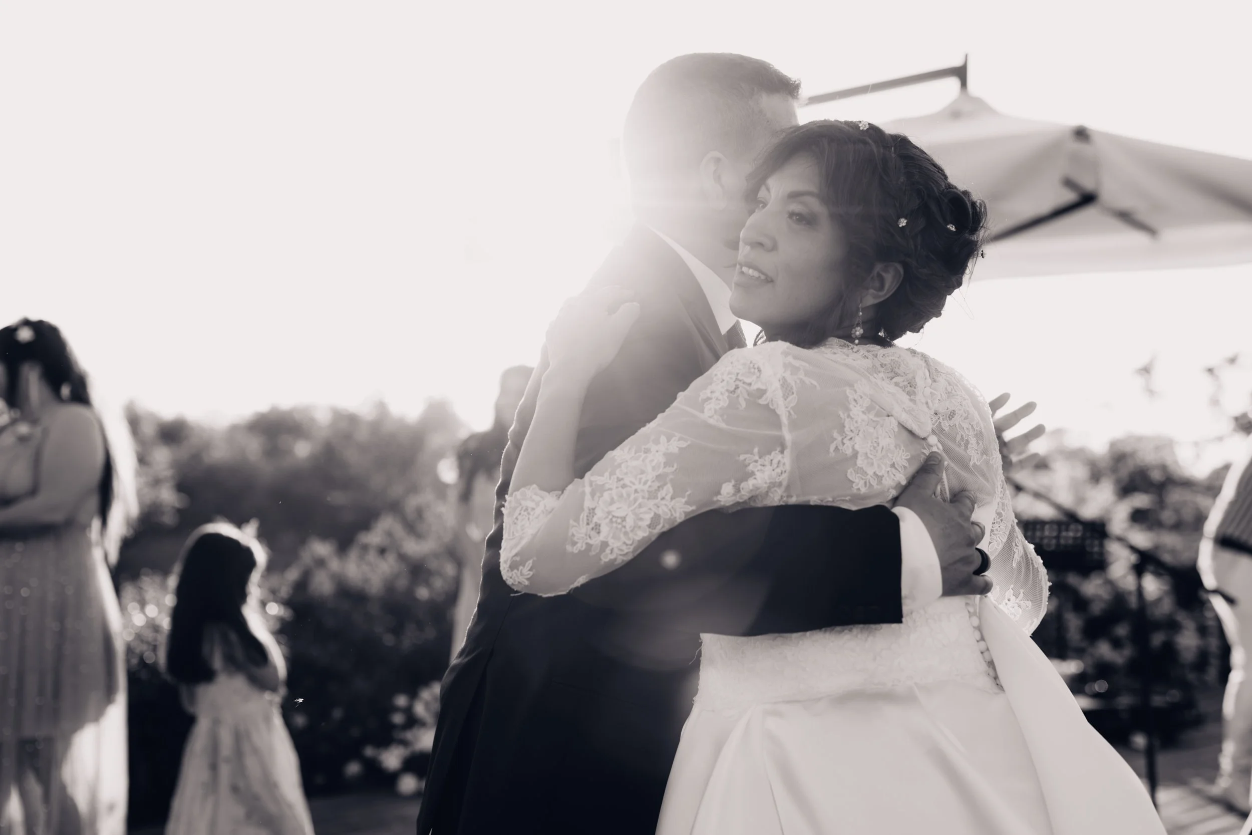 Immagine di una coppia di sposi che si abbracciano durante il loro matrimonio all'aperto, con altri ospiti sullo sfondo.