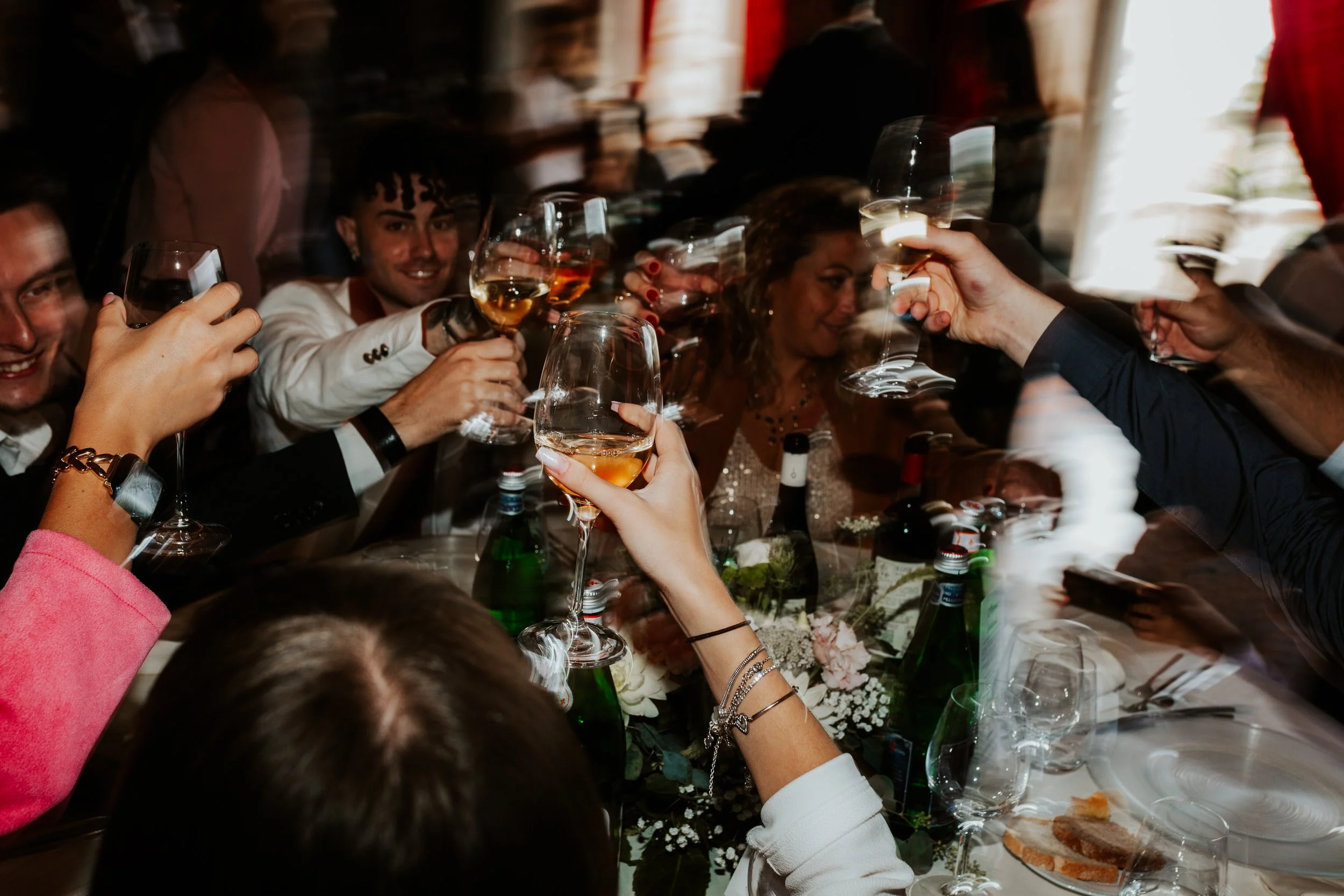Persone che brinda con calici di vino rosato durante una celebrazione, con un tavolo decorato con fiori e cibo.