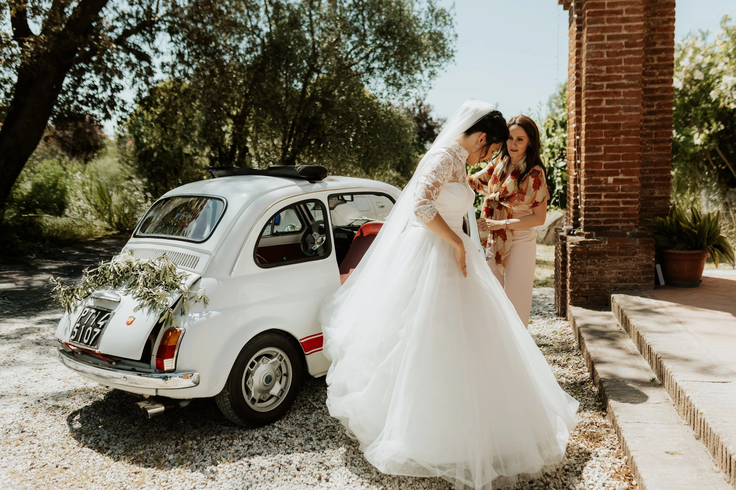 Una sposa in abito da matrimonio bianco con una donna accanto, vicino a una piccola auto vintage bianca decorata con rami e foglie, all'aperto in un giardino con alberi e un edificio di mattoni.