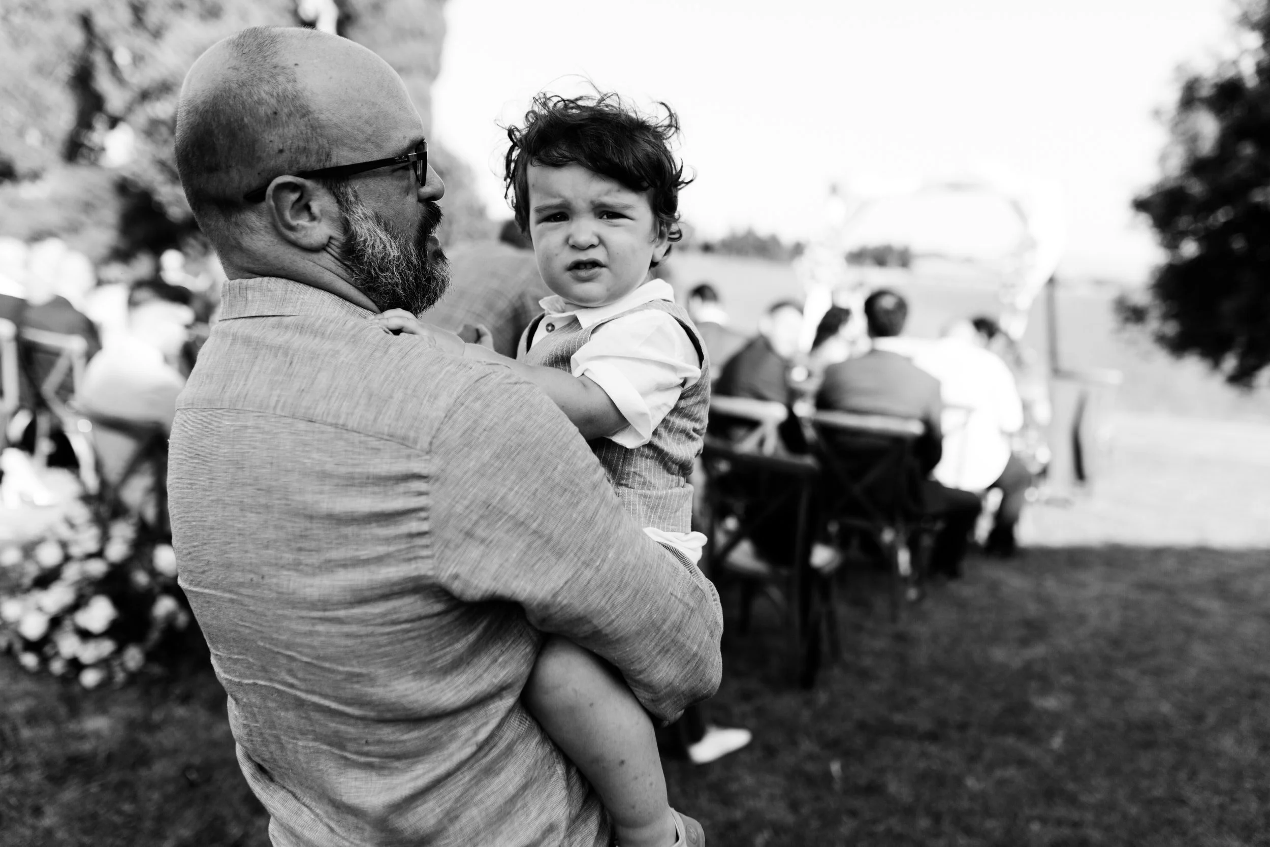 Uomo con barba e occhiali che tiene in braccio un bambino, scena di un evento all'aperto, persone sedute a tavoli sullo sfondo.