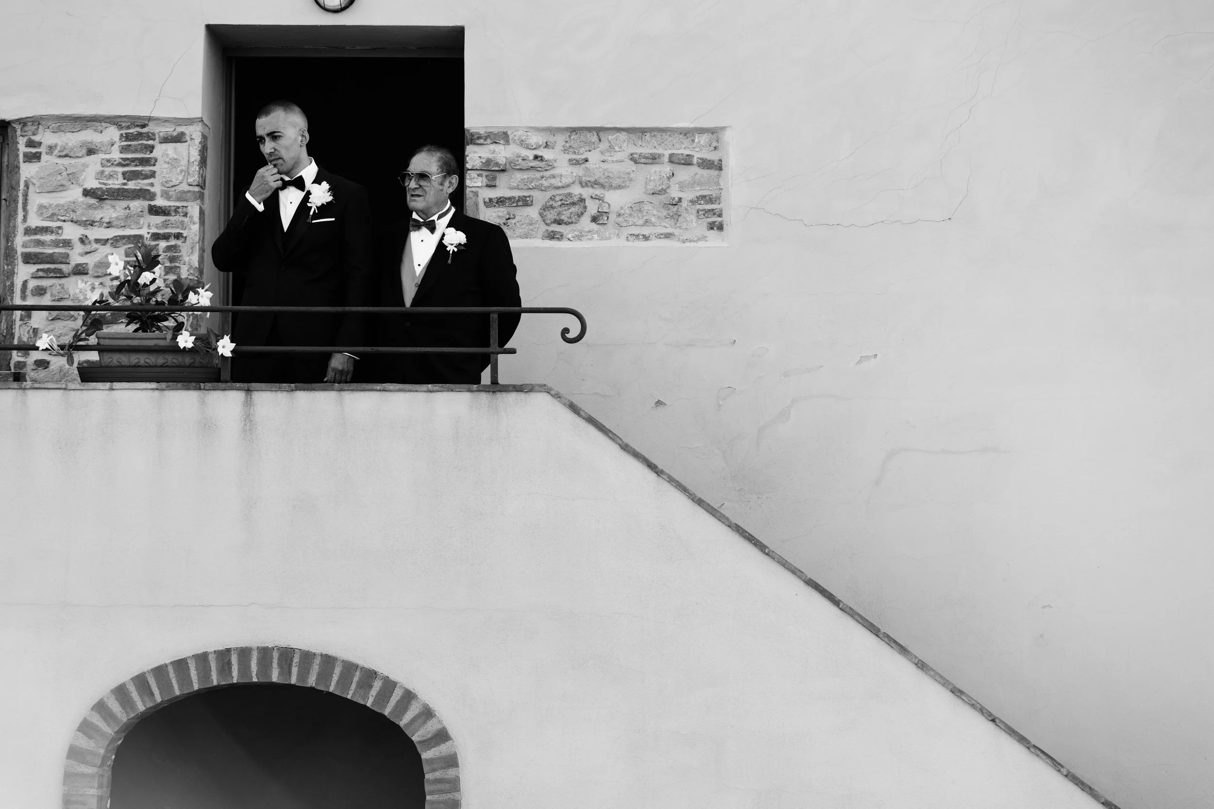 Due uomini eleganti in abiti formali con fiori sulla giacca, uno giovane e uno anziano, sono sul balcone di un edificio in stile classico con una parete bianca e un'arcata, in bianco e nero.