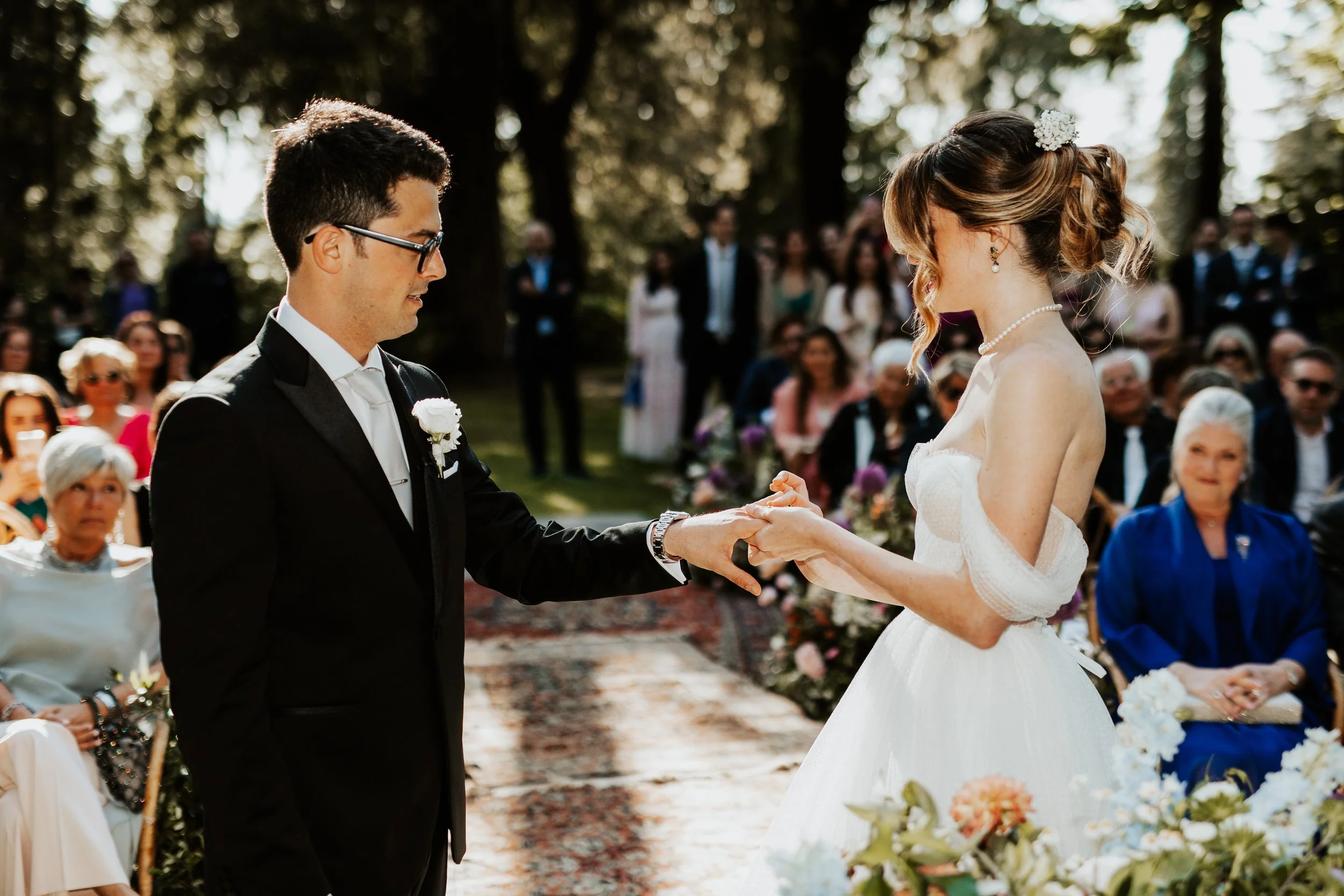 Una coppia di sposi durante il loro matrimonio all'aperto, lo sposo sta facendo una promessa e la sposa lo sta ascoltando, circondati da amici e familiari.