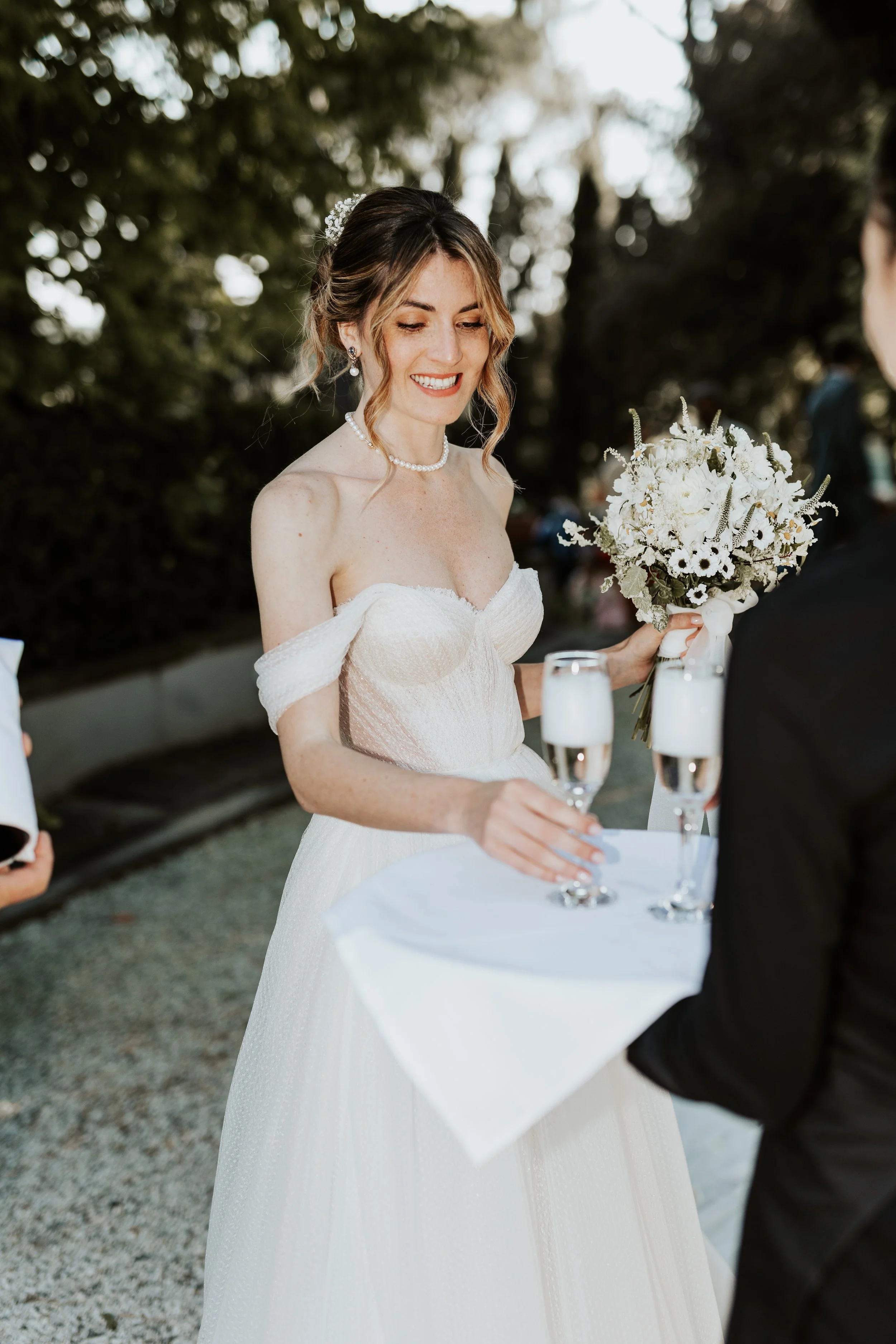 Sposa sorride durante un matrimonio, indossa abito bianco, collana di perle, e tiene un bouquet di fiori bianchi, mentre riceve un brindisi con due flute di champagne.