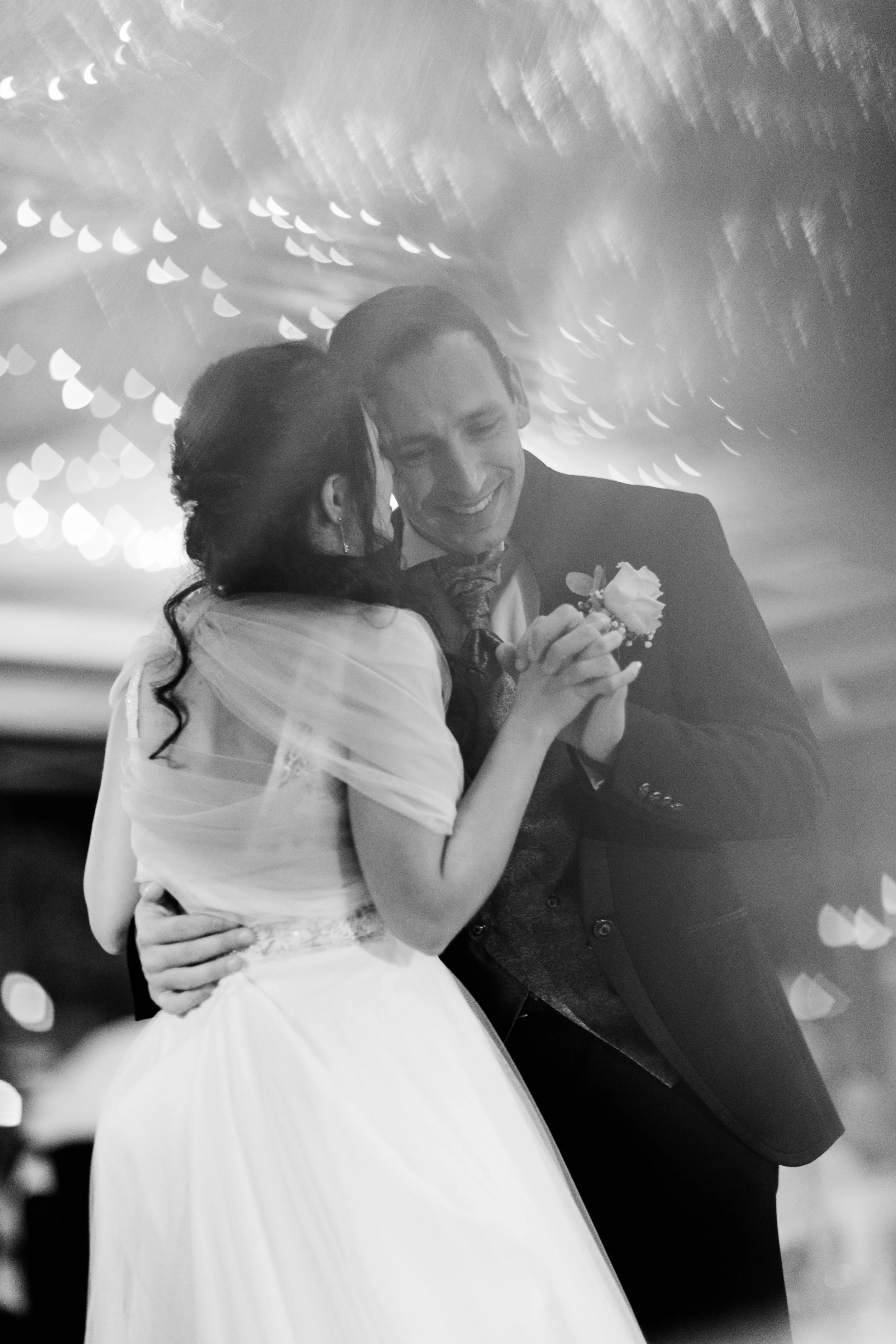 Due persone in abiti da sposo si tengono per mano e sorridono, probabilmente durante un ballo di nozze, in un ambiente con luci soffuse e decorazioni di festa.