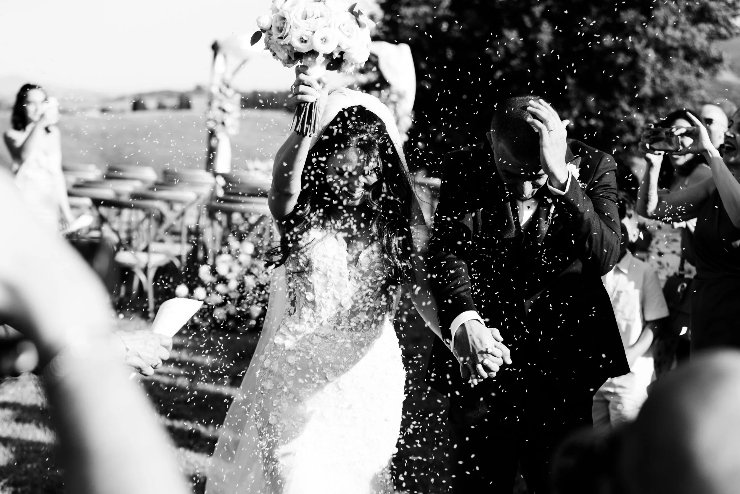 Una coppia di sposi, donna in abito da sposa e uomo in abito formale, si tengono per mano durante il loro matrimonio esterno, circondati da amici e parenti che lanciano coriandoli.